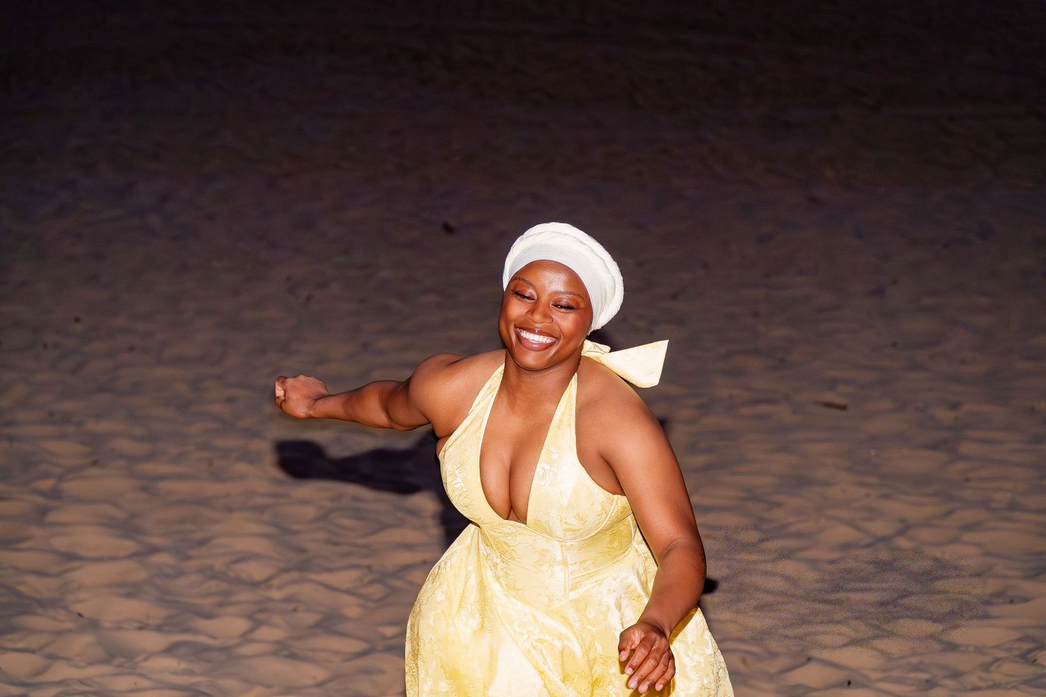 A joyful woman in a yellow halter dress and white head wrap dances barefoot on a sandy beach at night.