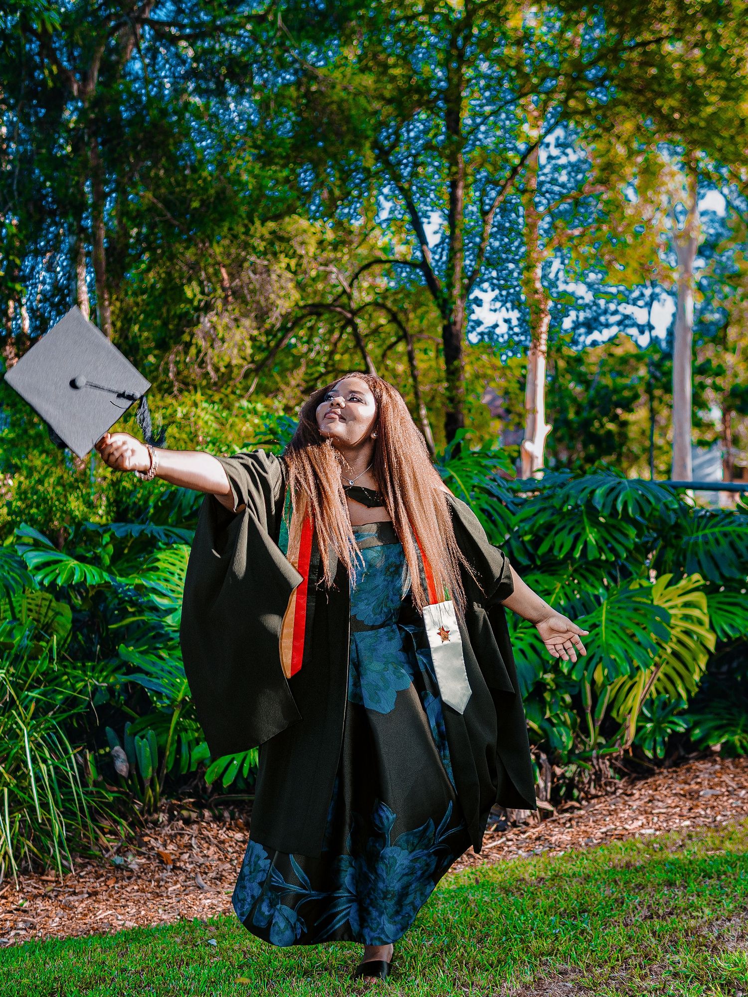Elated graduate in black gown twirls with mortarboard cap outstretched in lush green garden setting.