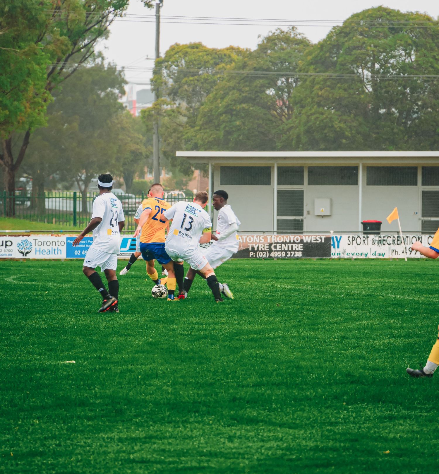 Soccer players compete for the ball during an outdoor match on a green grass field with trees and a clubhouse in the background.