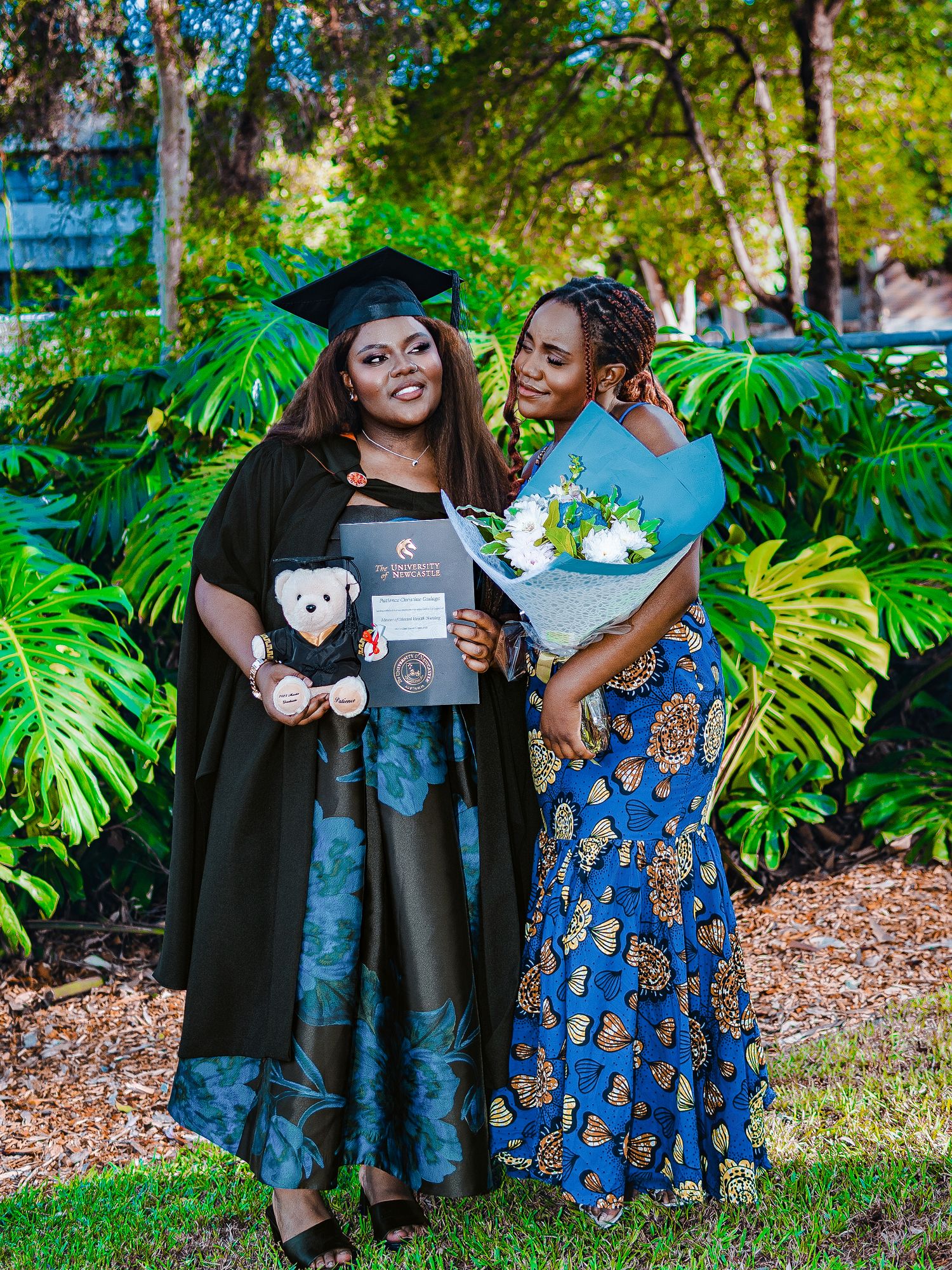 Two women pose together, one in graduation cap and gown holding diploma and teddy bear.