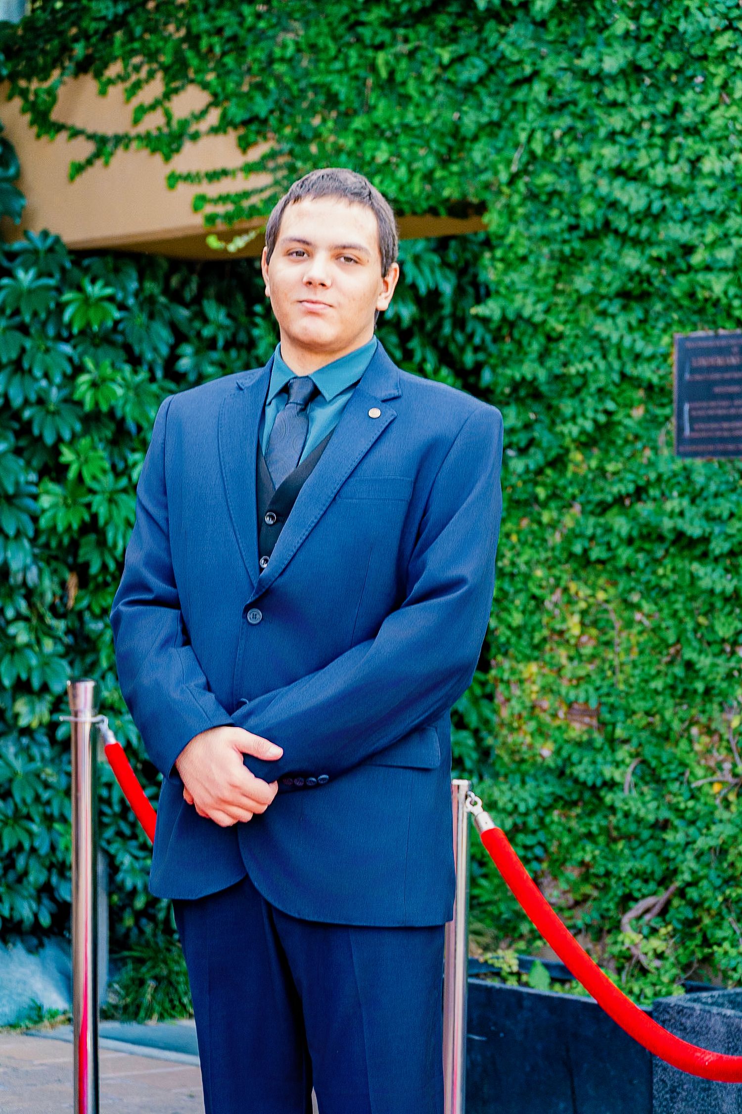 Young man in navy blue suit with green tie standing at formal event entrance with red rope barriers.