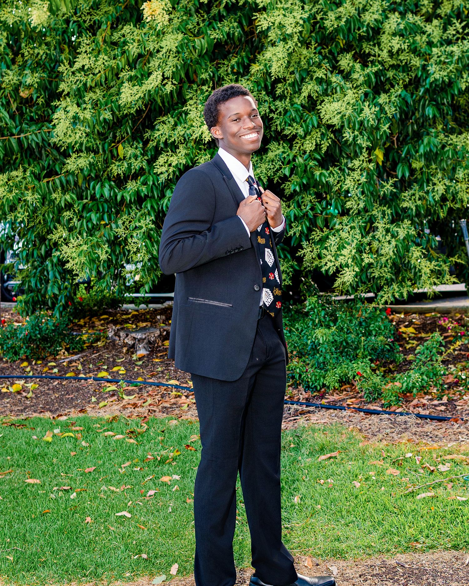Young man in black suit smiling outdoors, posing confidently in front of lush green hedges on a sunny day.