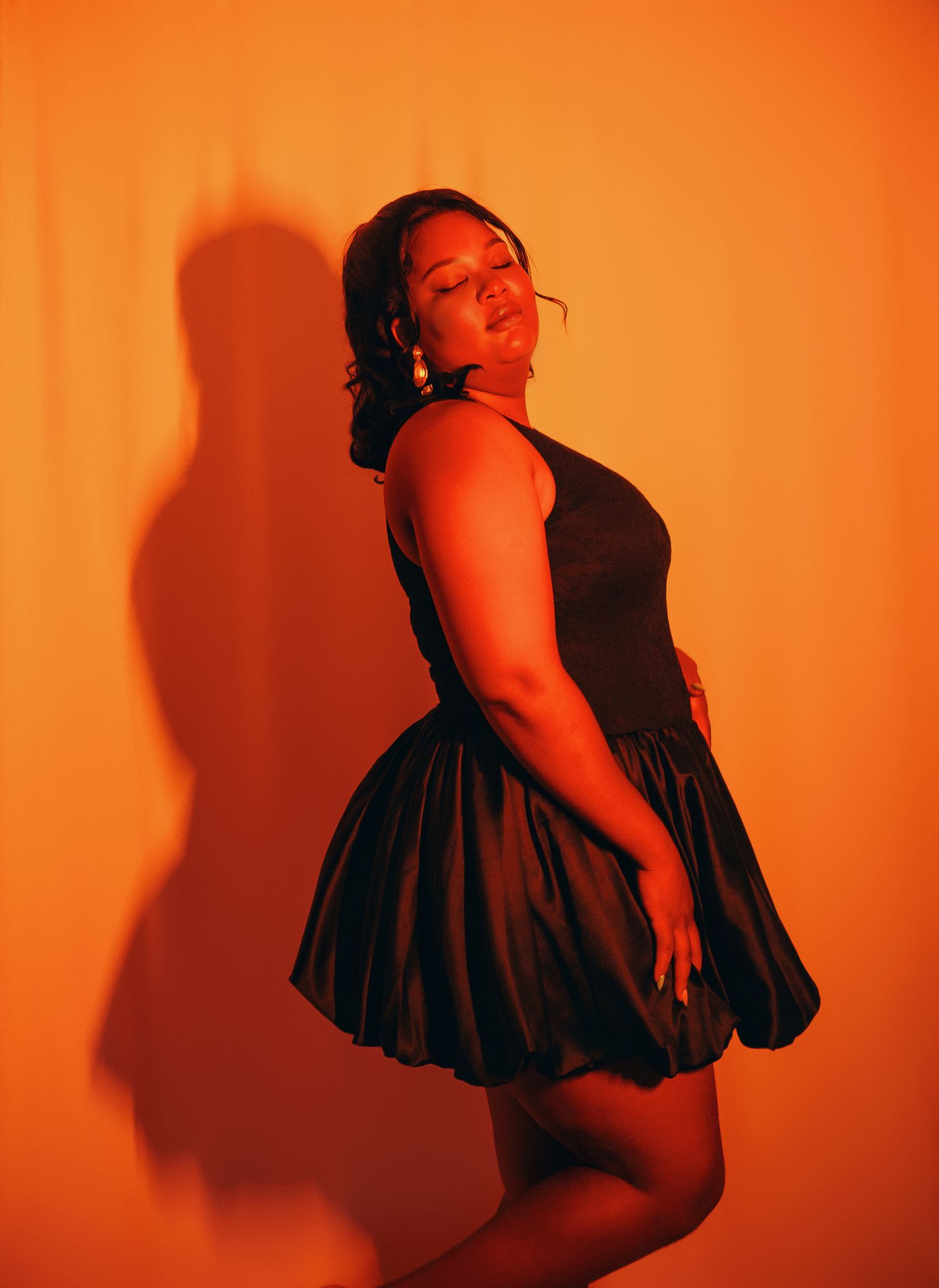 Woman in black satin bubble dress posing sideways in orange studio.