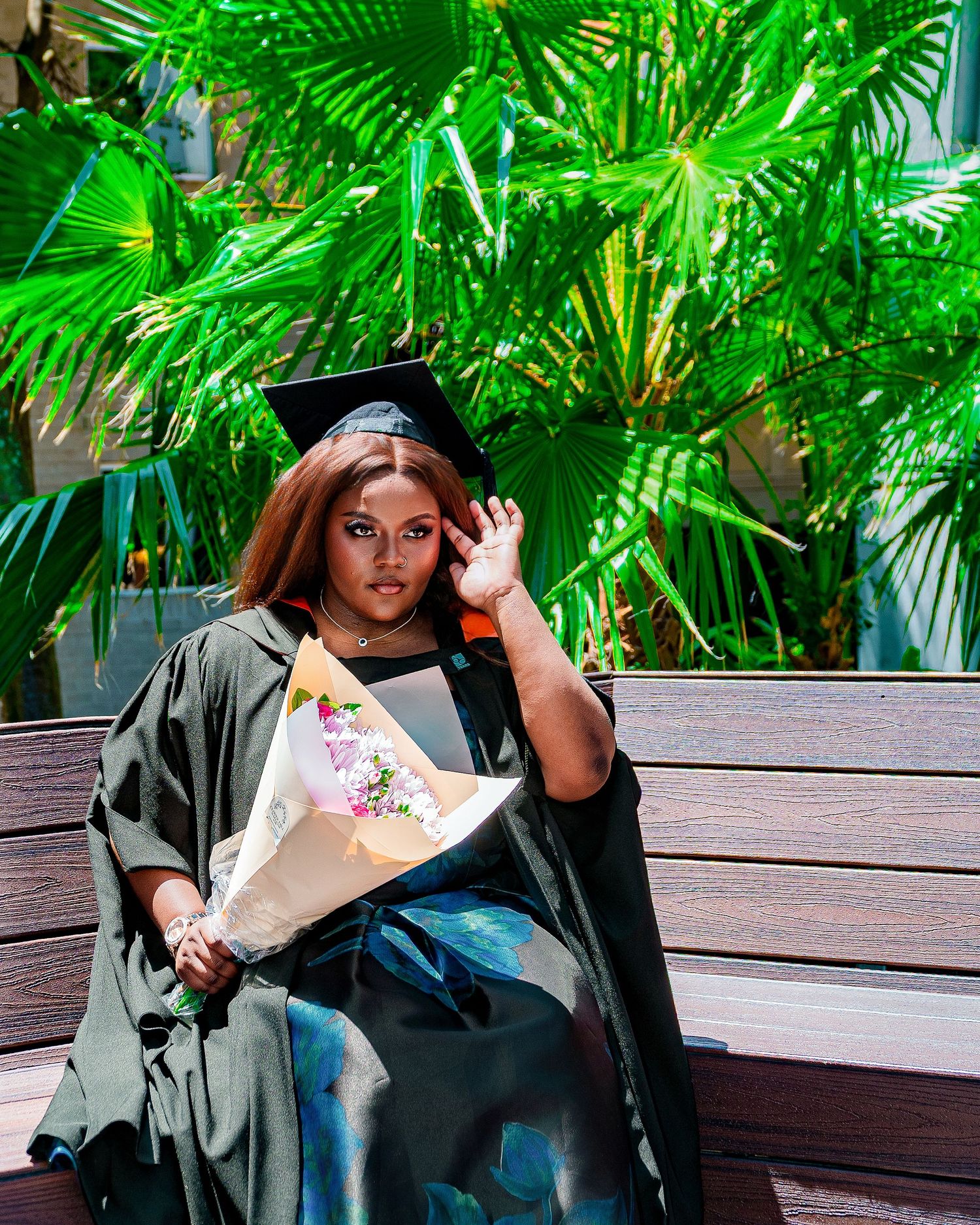 A young woman in graduation cap and gown holds a bouquet of flowers while seated on a bench surrounded by tropical palms.
