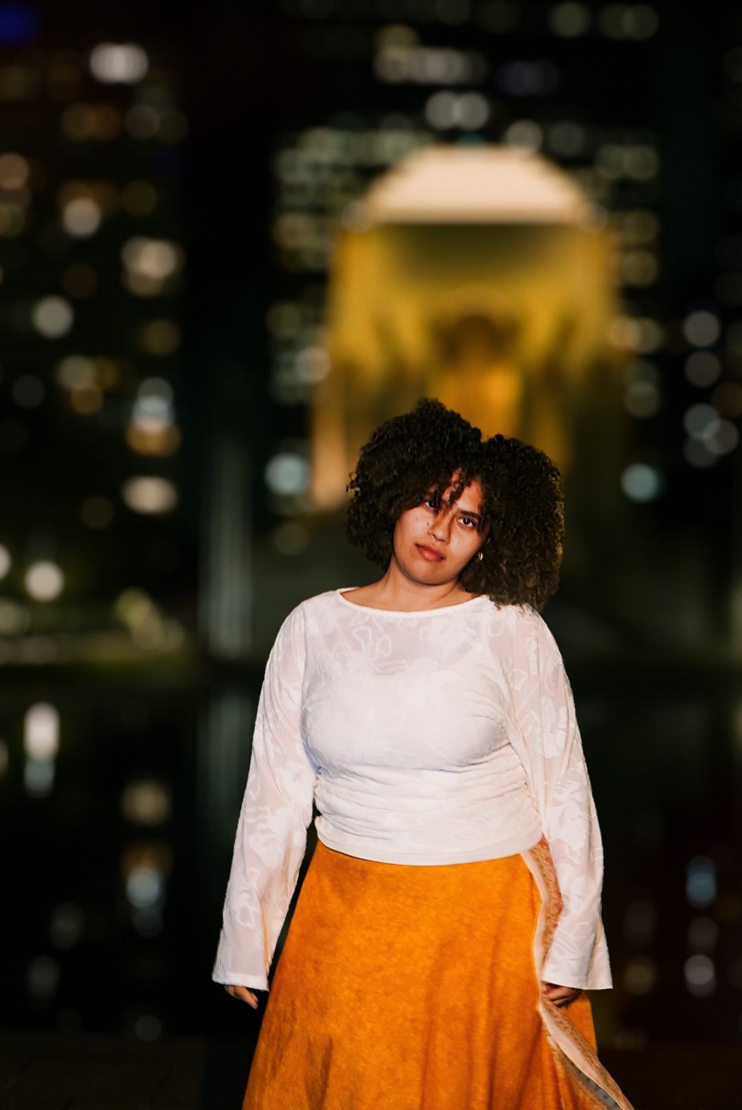 Young woman wearing white blouse and orange skirt standing outdoors at night with bokeh city lights background.