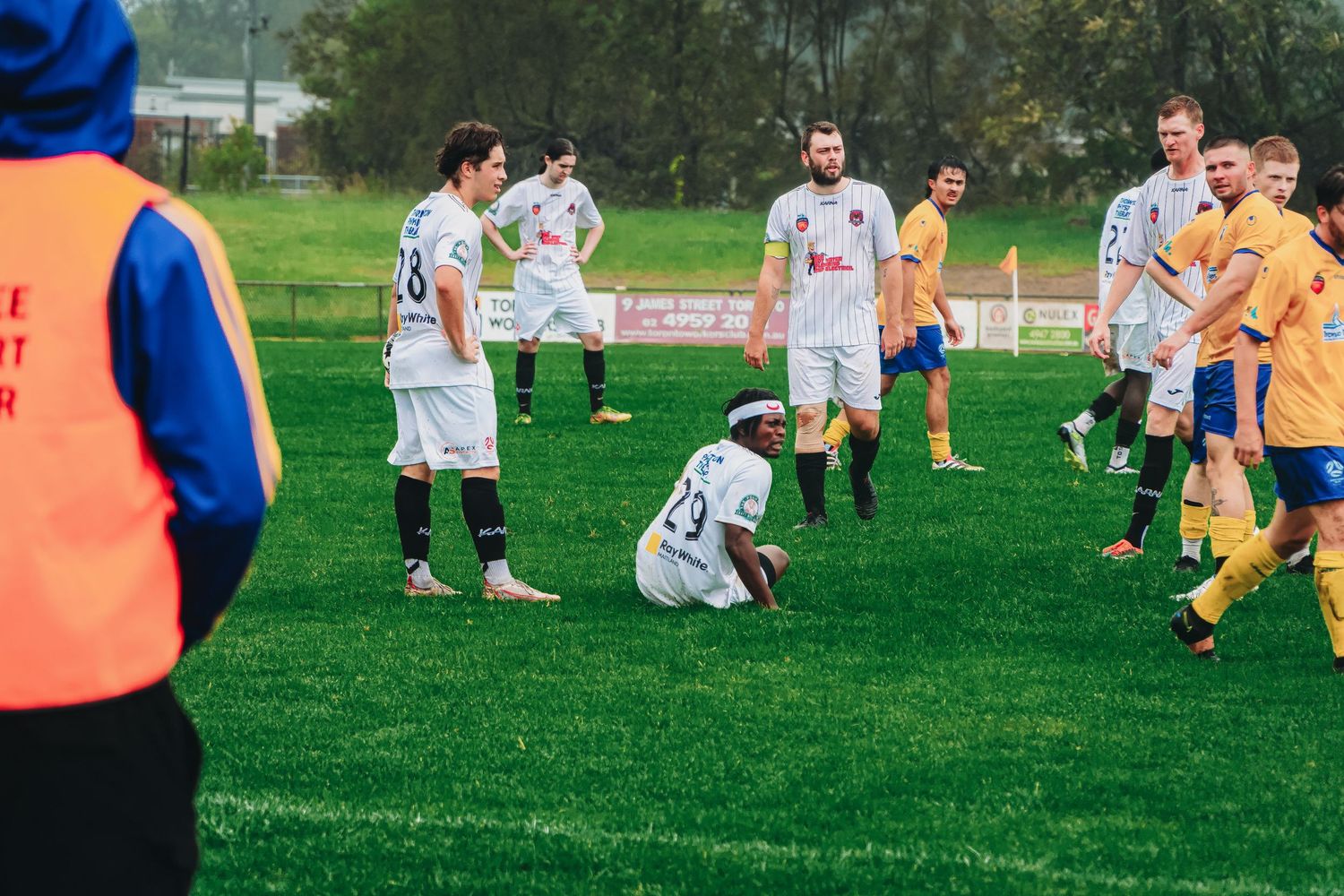 Soccer player sits on green field during a break in play while teammates stand nearby.