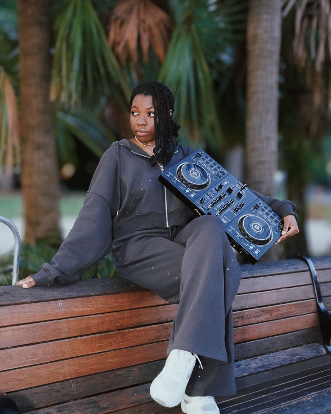 Young woman in gray tracksuit holds DJ controller while sitting on wooden bench outdoors.