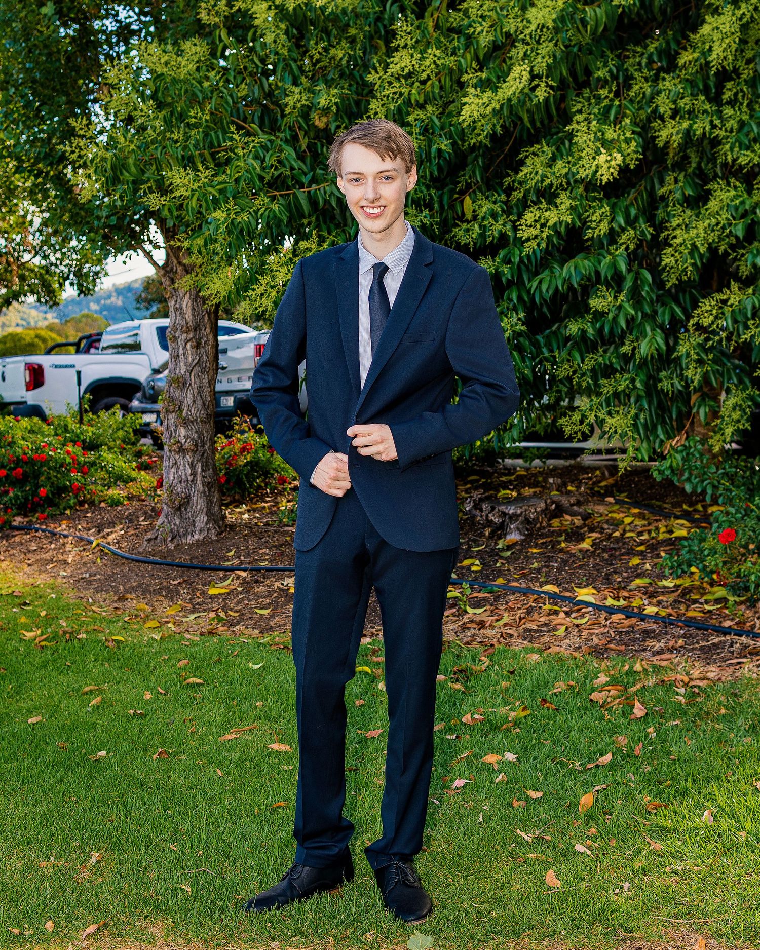 Young man in navy blue suit and tie posing outdoors in a garden setting with green trees and red flowers.