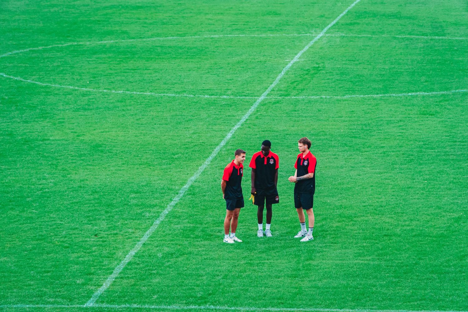 Three soccer players in red and black uniforms stand on a lush green field discussing strategy near a white boundary line.