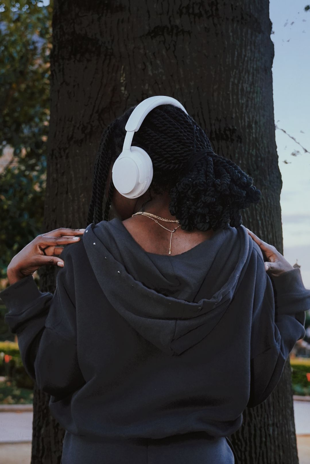 Woman with white headphones standing by tree, rear view.
