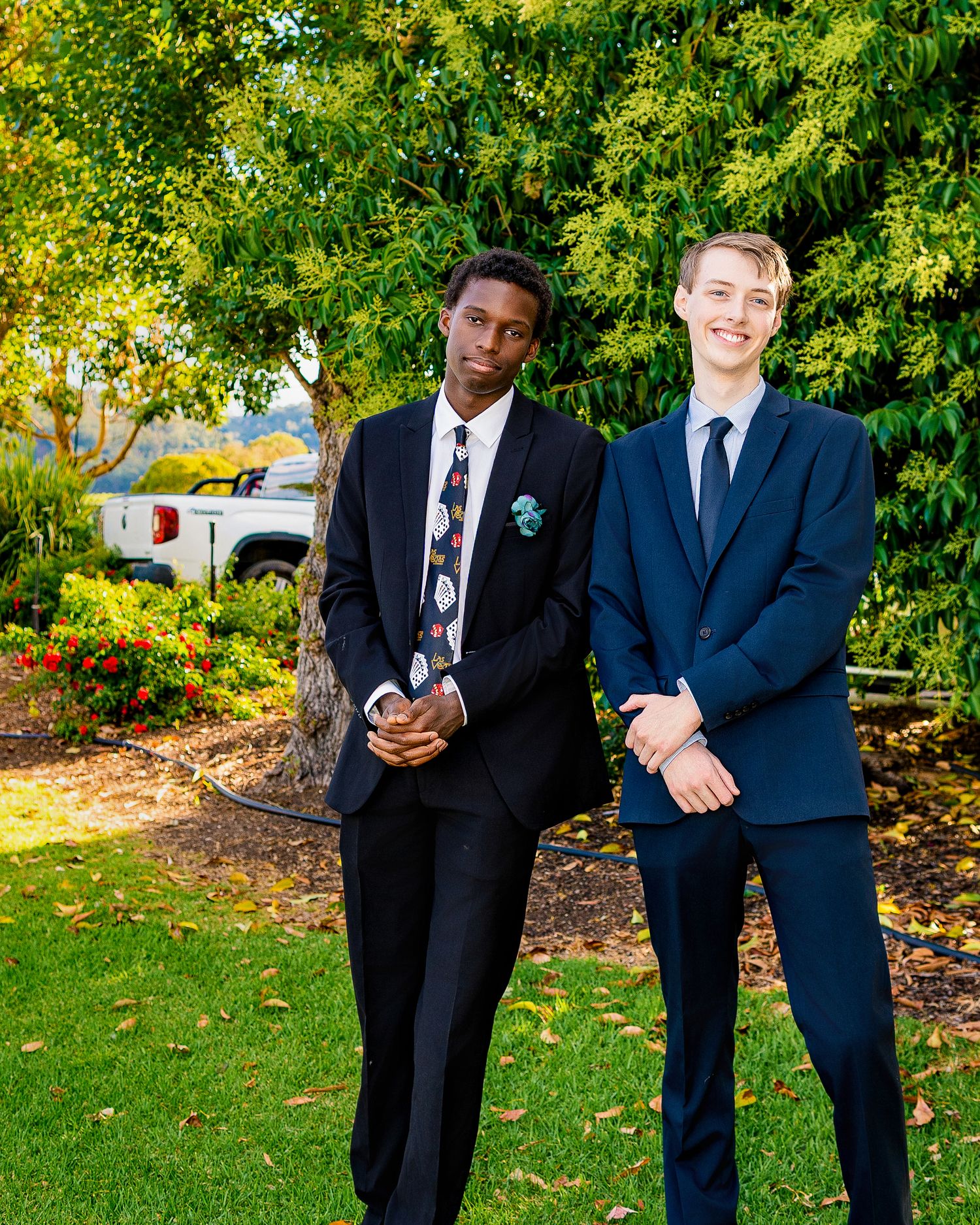 Two young men in formal black suits standing on green grass outdoors near trees and a white truck.
