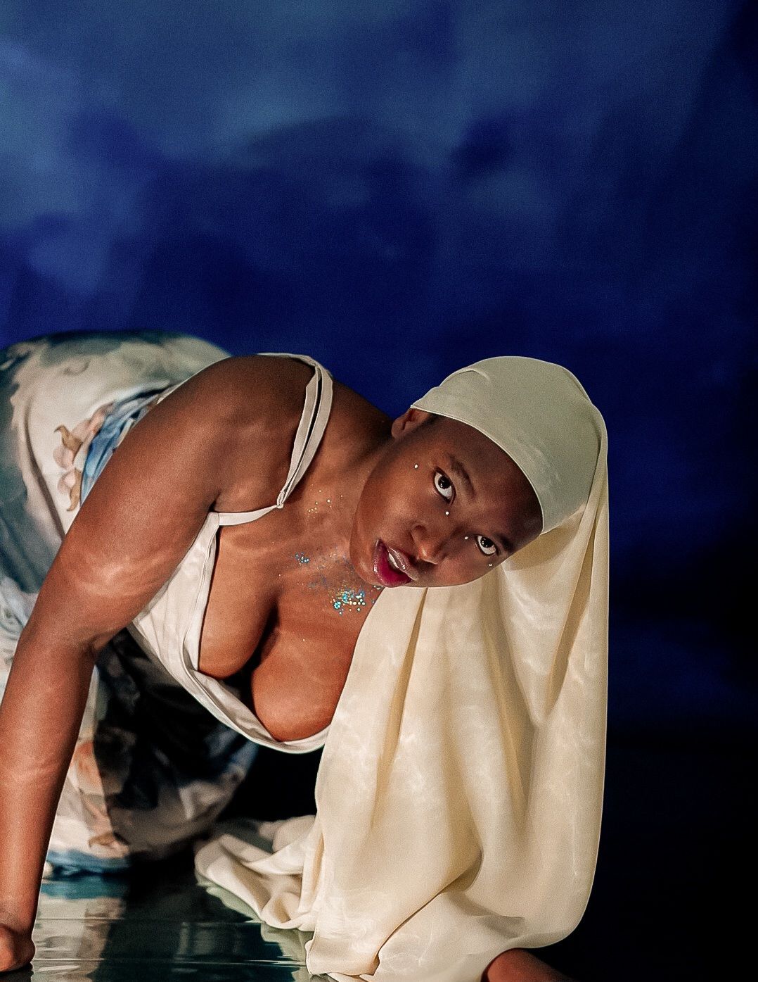 A woman wearing a cream hijab and floral outfit poses dramatically against a dark blue background.