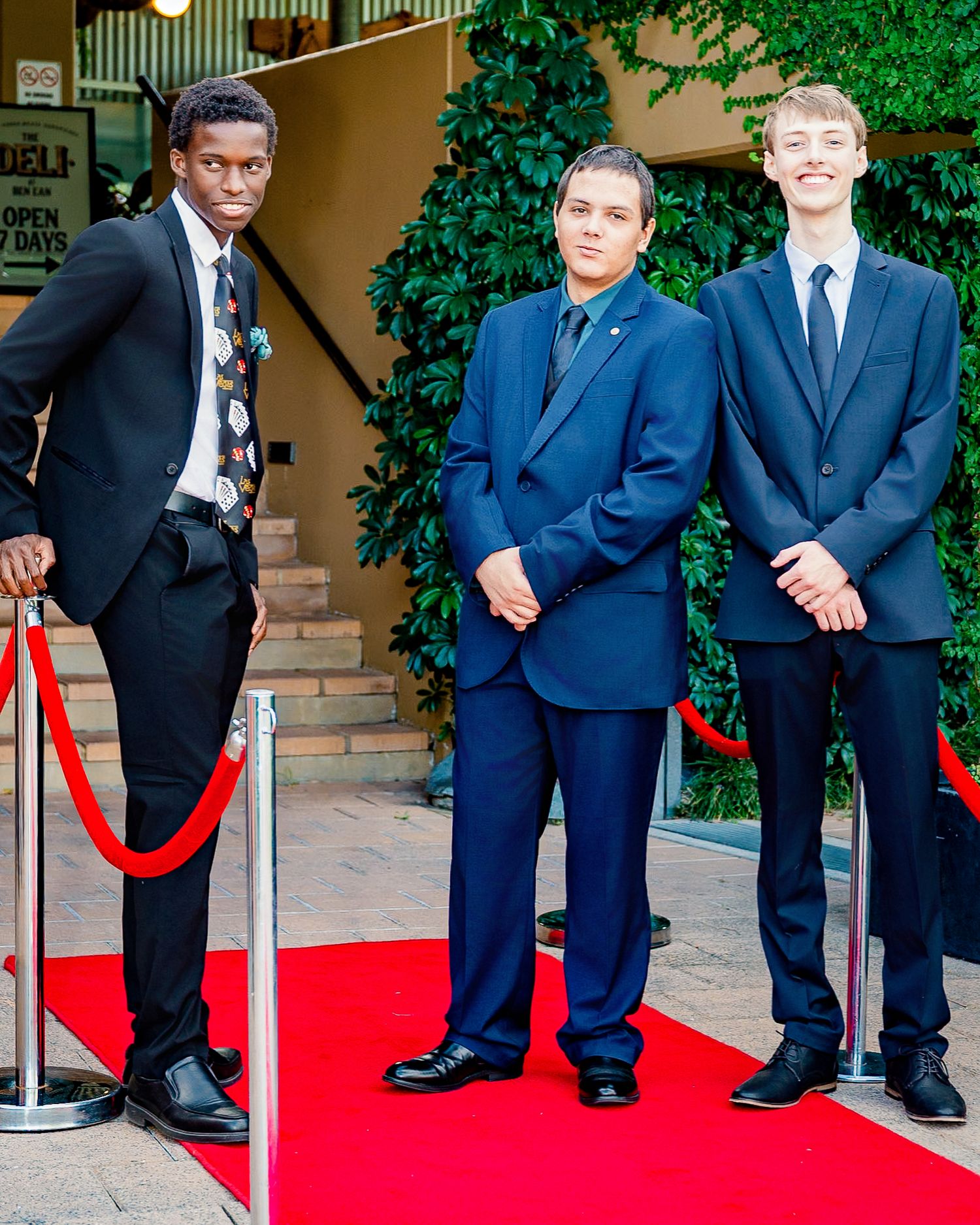 Three young men in formal suits stand on a red carpet at an elegant event venue.