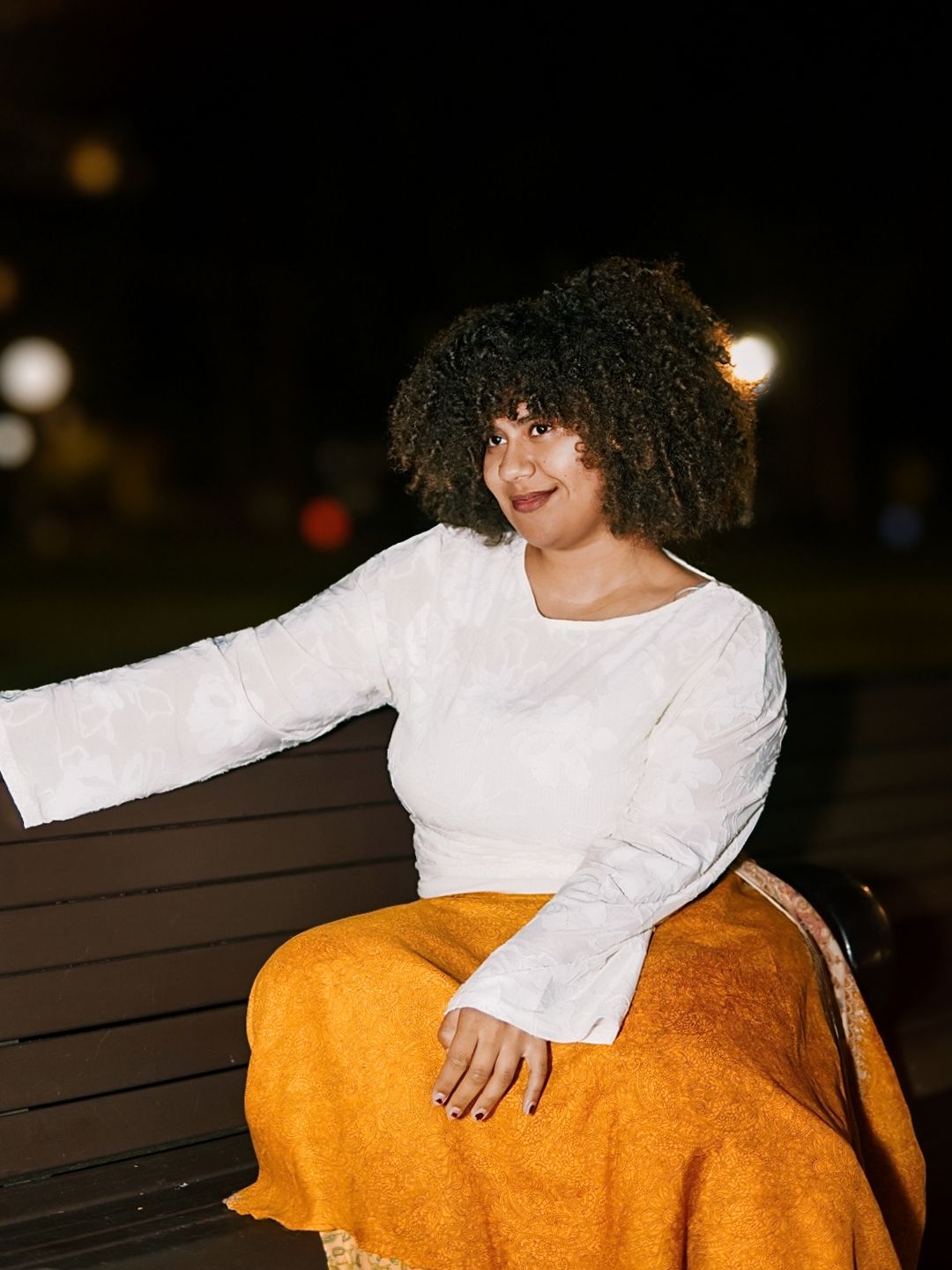 Curly-haired woman in white long-sleeve top and orange skirt poses on a bench at night.