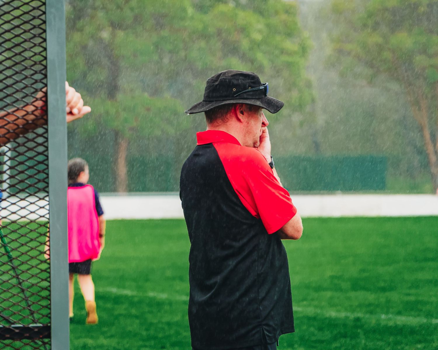 A coach in a red and black shirt and black hat watches a rainy soccer match from the sideline.