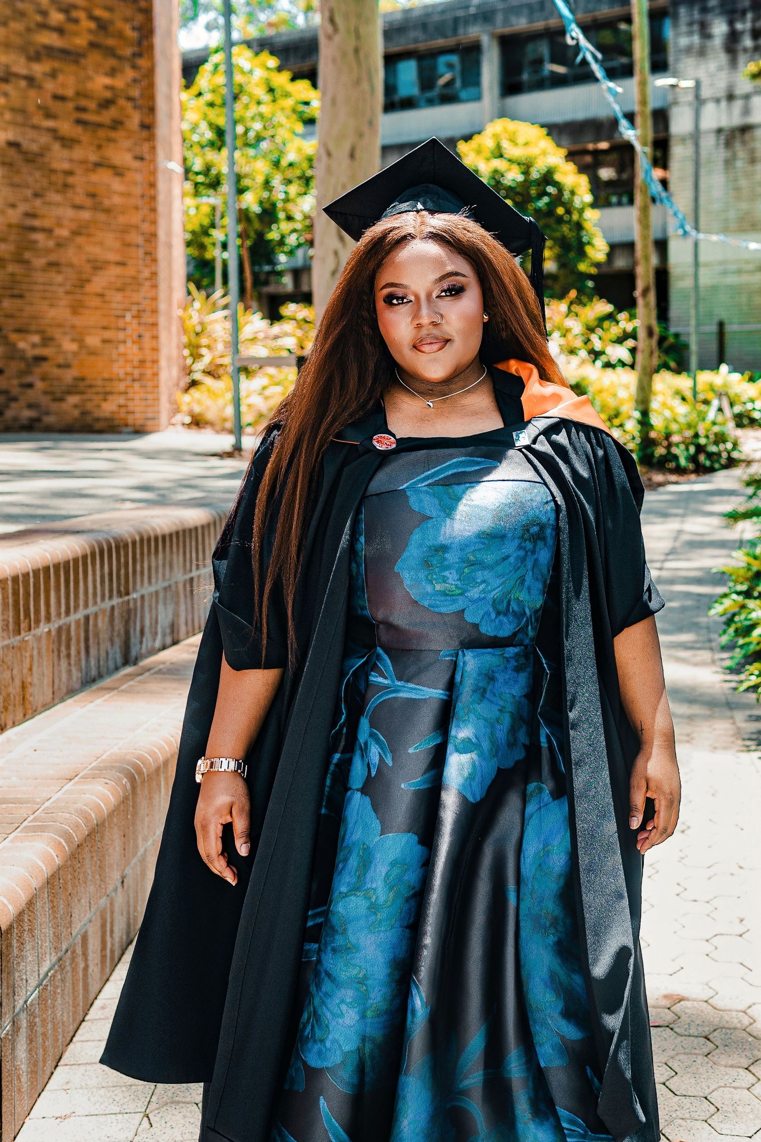 Graduate in black cap and gown over floral blue dress poses outdoors at university campus.