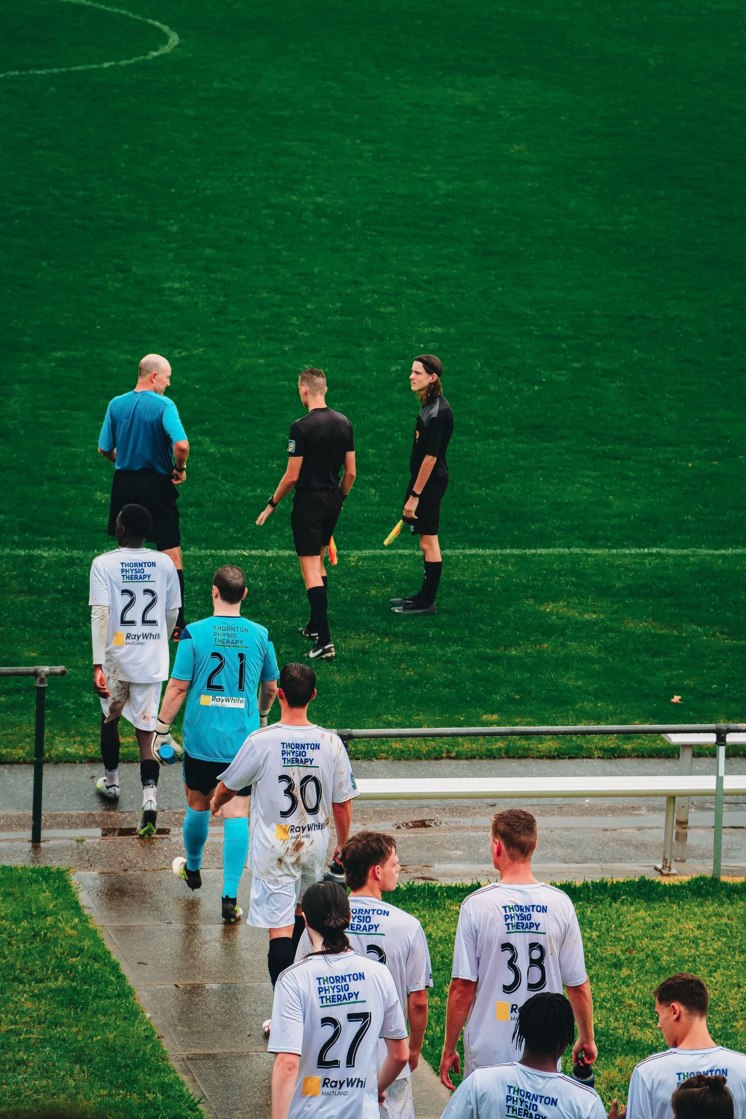 Soccer players in white jerseys with numbers walk onto a green field alongside referees in black uniforms.