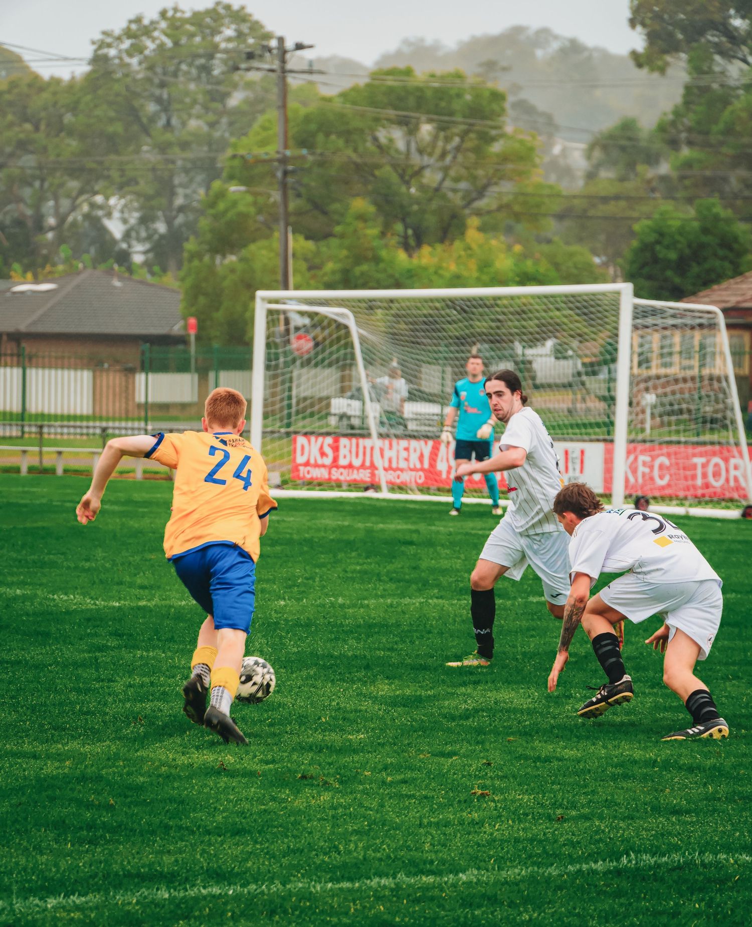 Soccer players compete on green field, player in yellow jersey number 24 dribbles ball against white-uniformed defenders.