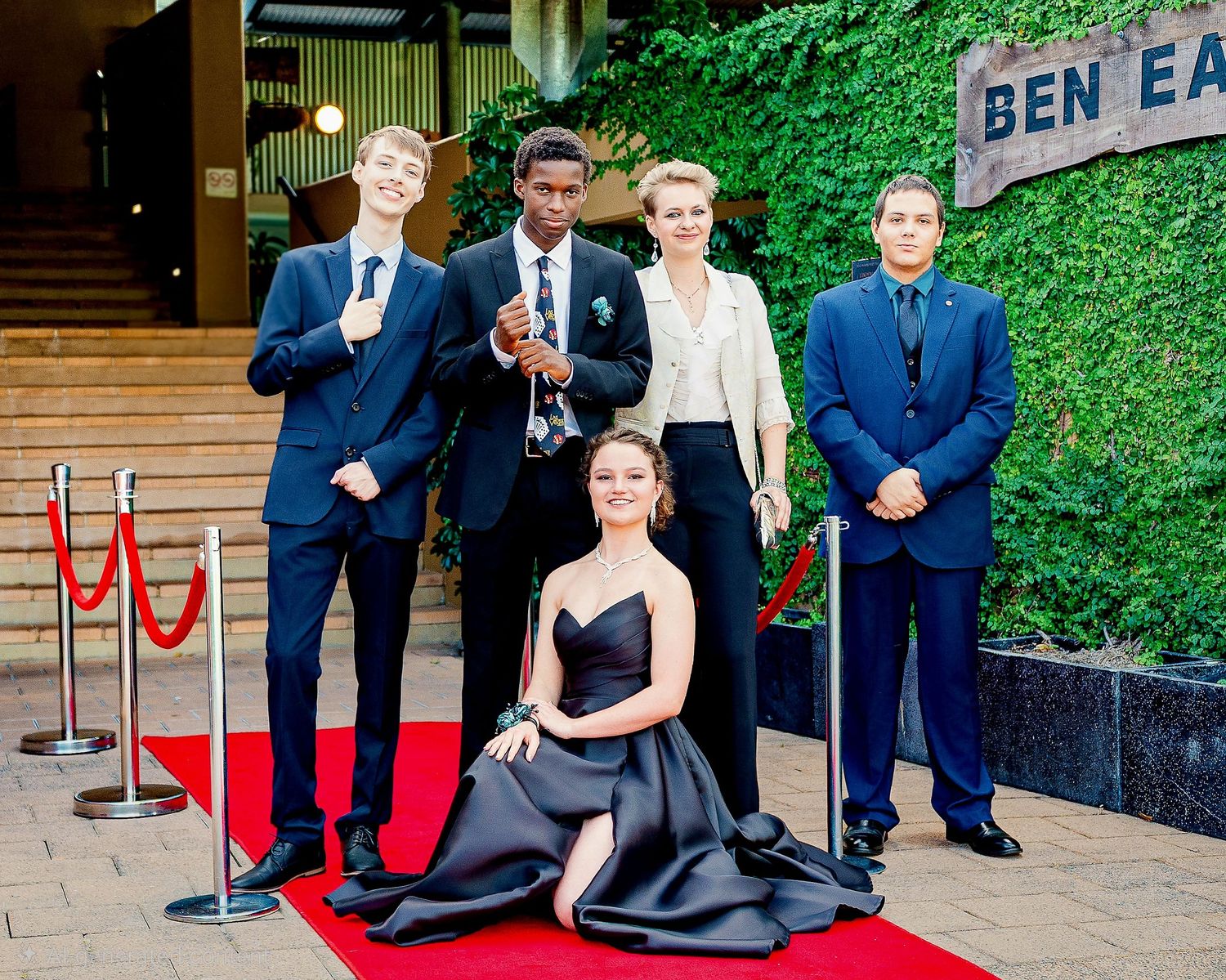 Five elegantly dressed teens pose on a red carpet at a formal prom event in front of a green ivy wall.