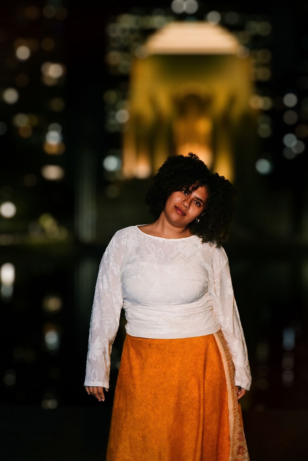 Woman in white lace top and orange skirt posing confidently at night with blurred city lights behind her.
