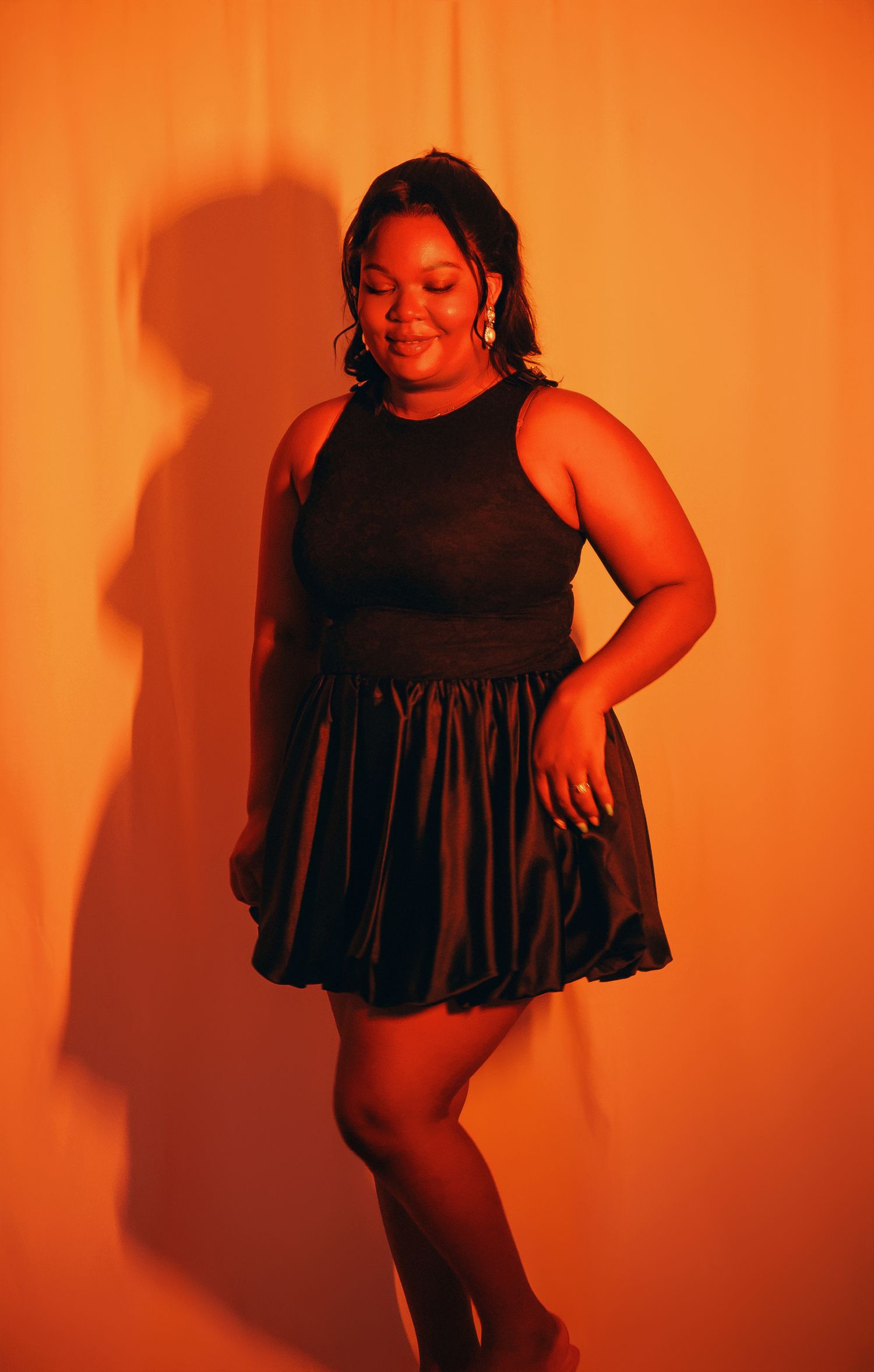 Woman in black mini dress posing against orange backdrop.