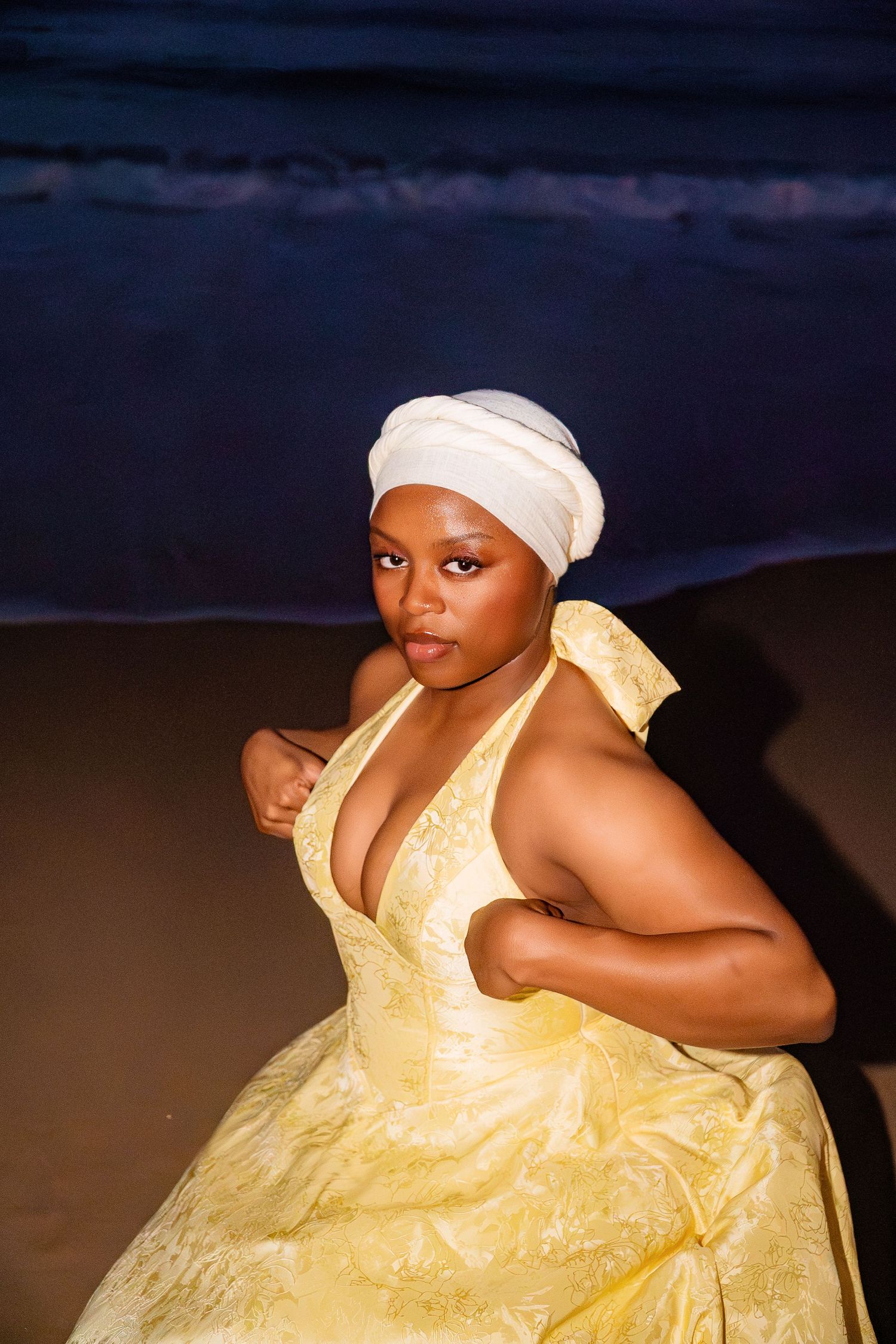 Woman in yellow halter dress and white head wrap poses confidently on a beach at night.