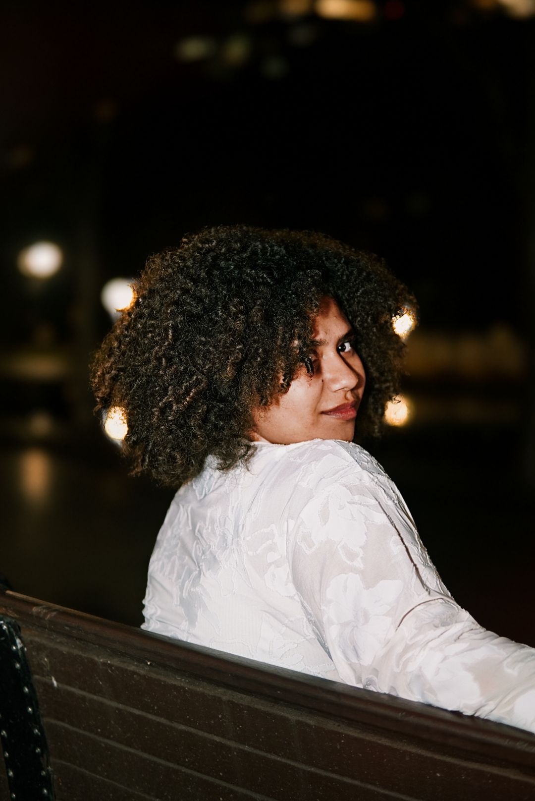 Woman with voluminous curly hair in white top glances back over shoulder on bench at night.