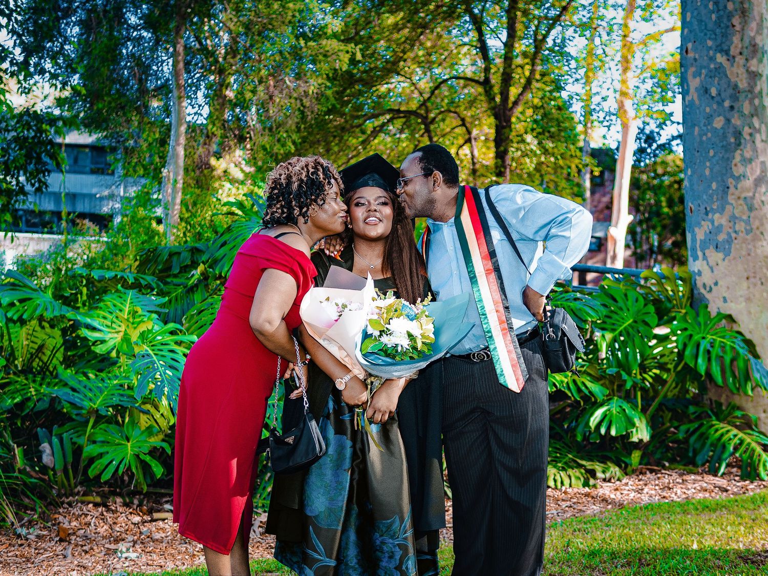 Graduate in cap and gown receives kisses from proud parents while holding bouquet outdoors.