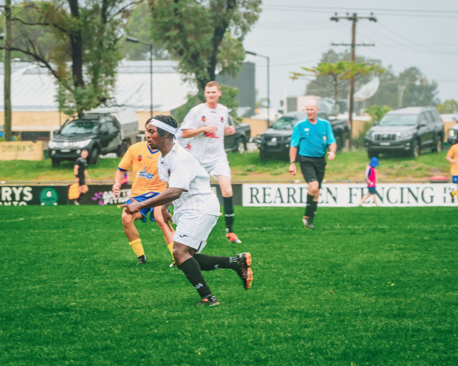 Soccer player in white uniform dribbling ball on green field during competitive match with referee and spectators.