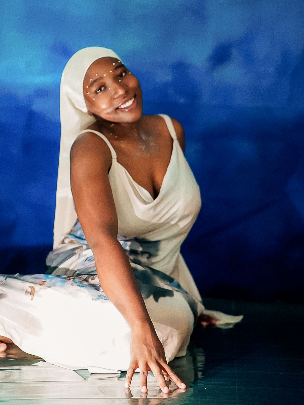 A smiling woman in a white hijab and floral dress poses gracefully on the floor against a blue background.