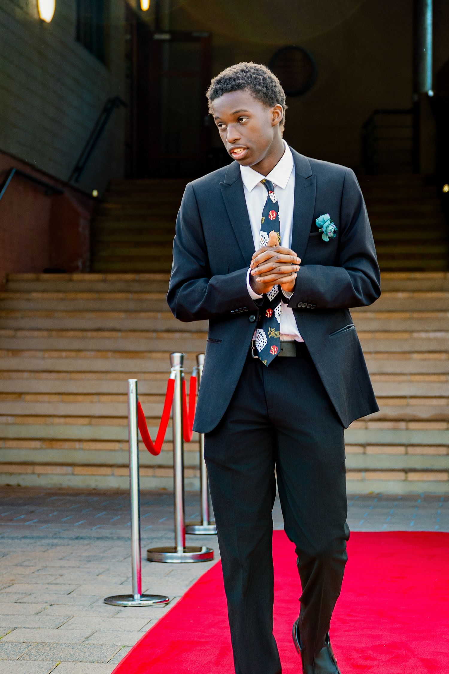 Young man in black suit with colorful tie stands on red carpet near velvet rope barriers.