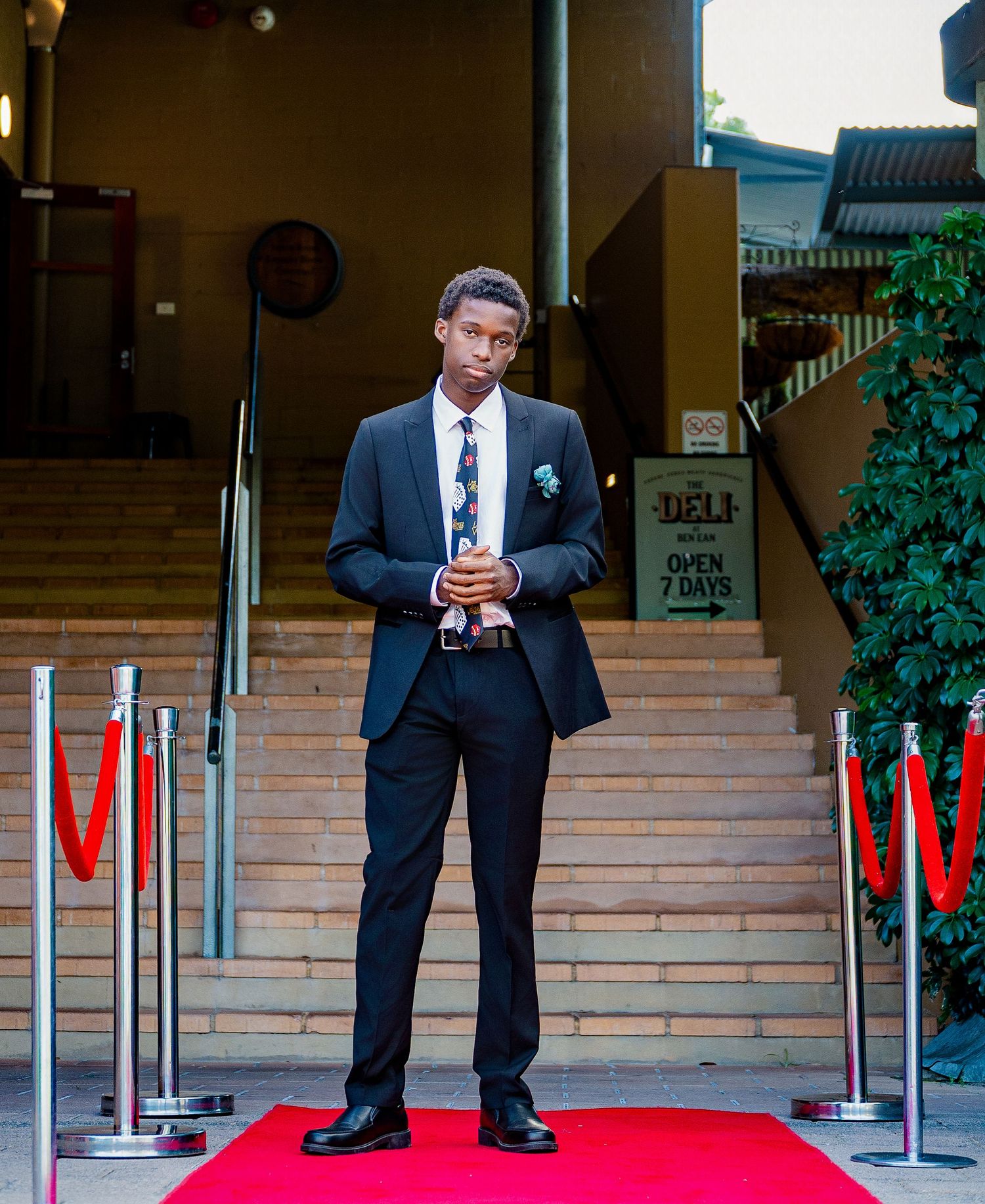Young man in black suit and floral tie stands on red carpet outside a deli entrance.