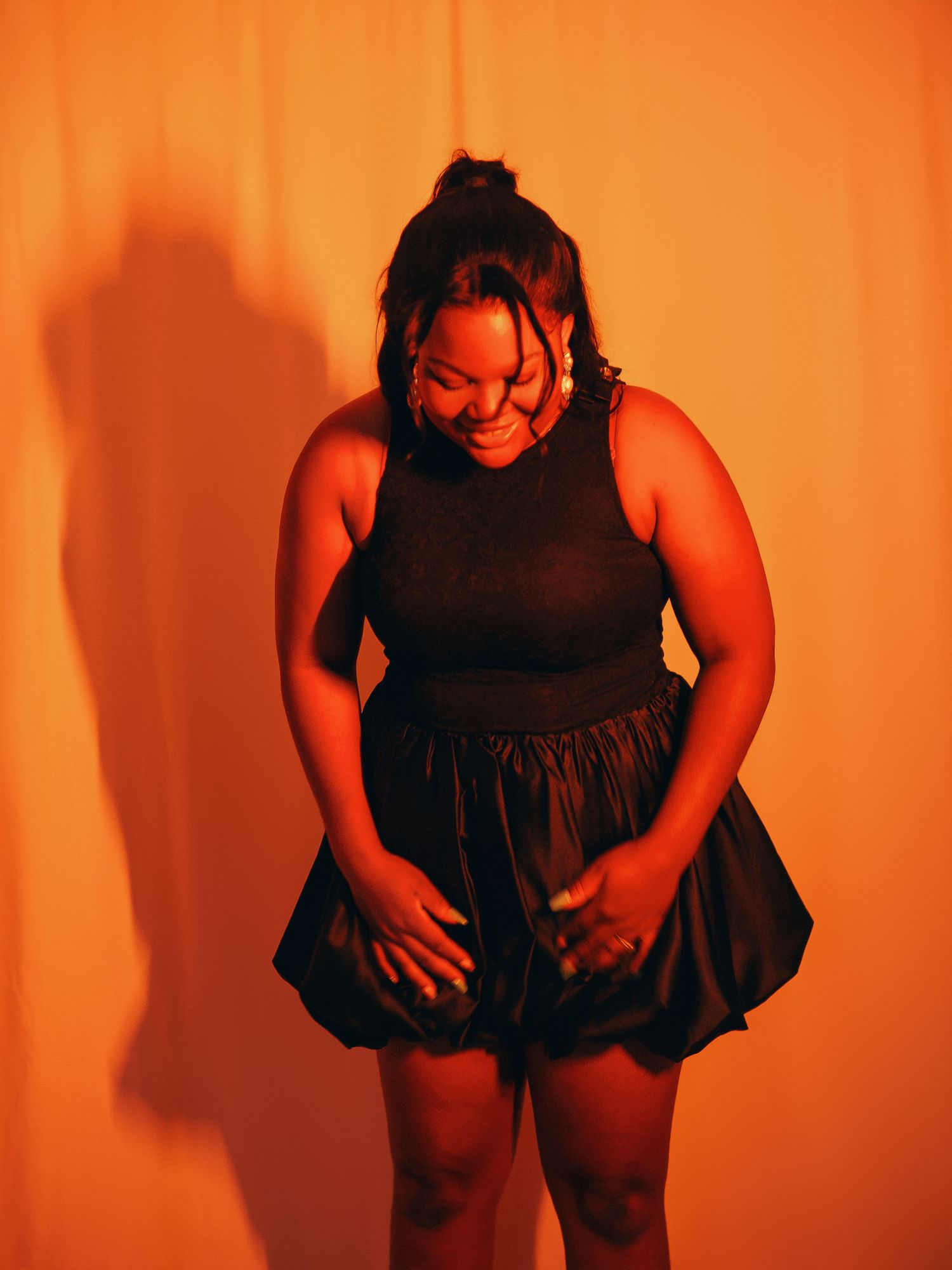 A plus-size woman in a black mini dress poses confidently under warm orange lighting against a draped backdrop.
