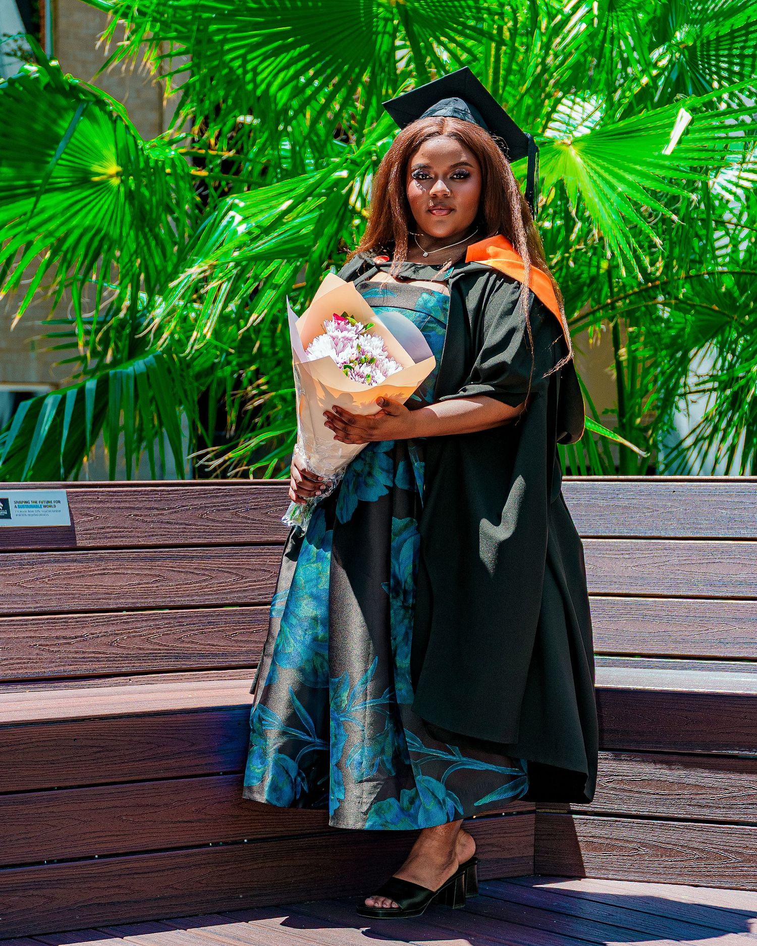 Graduate in black cap and gown holds bouquet standing before lush tropical palm leaves on bench.