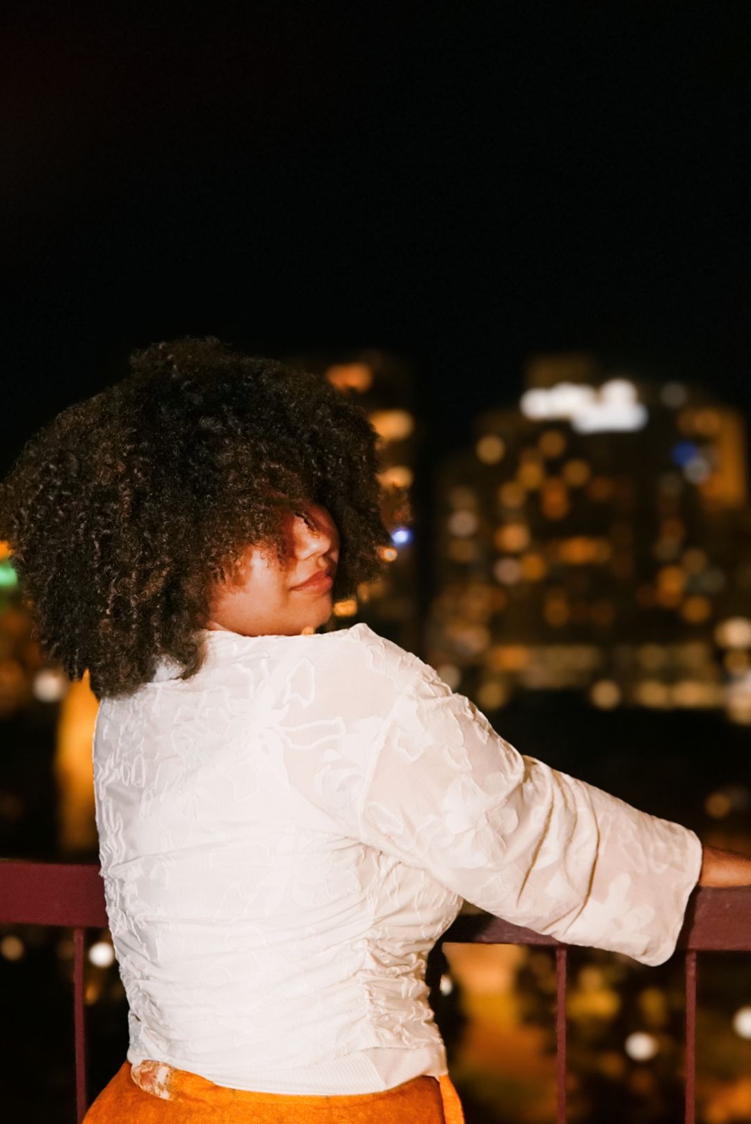 Back view of woman with curly hair in white top and orange skirt overlooking glowing city lights at night.