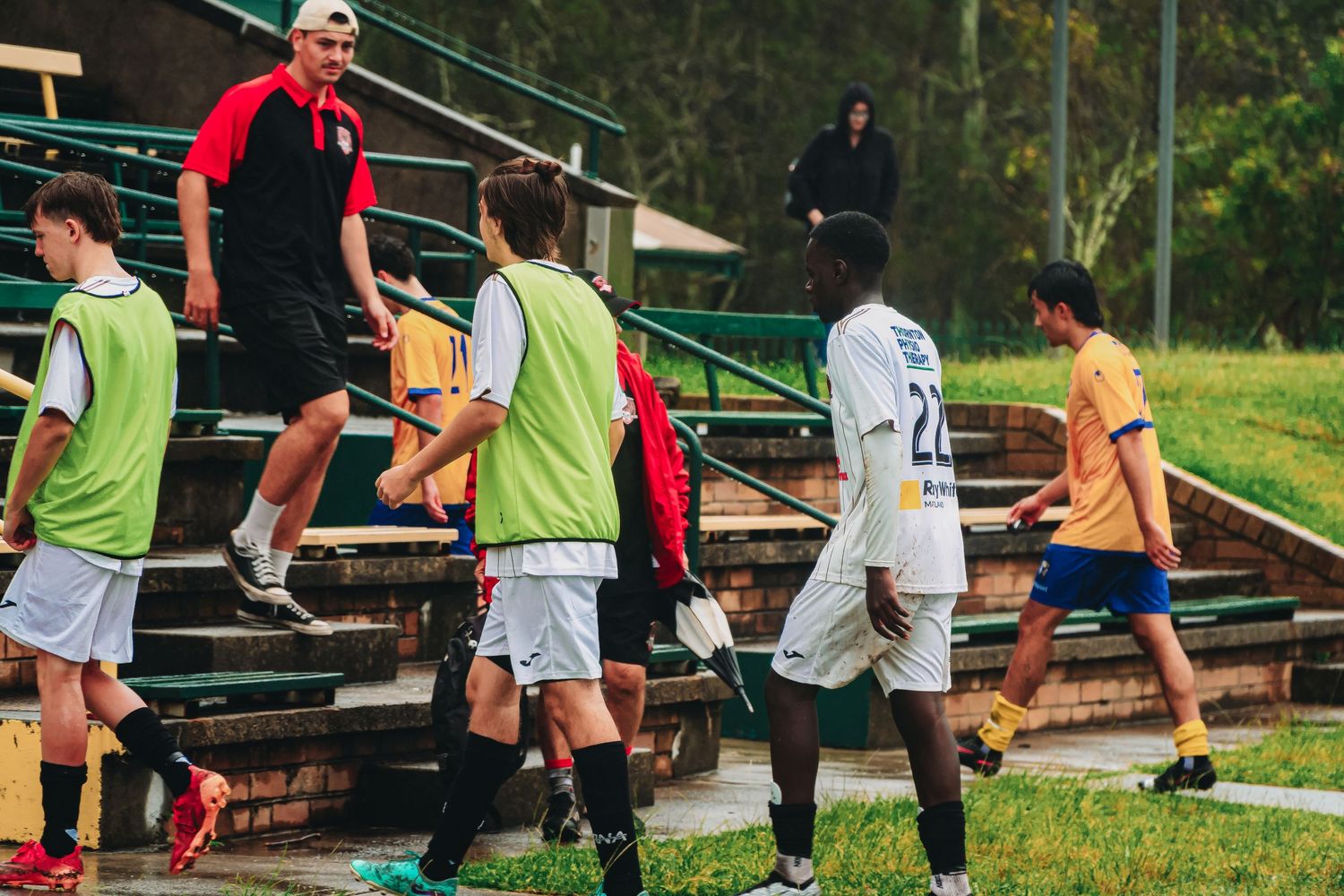 Youth soccer players in colorful bibs walk near stadium bleachers with a coach during training session.