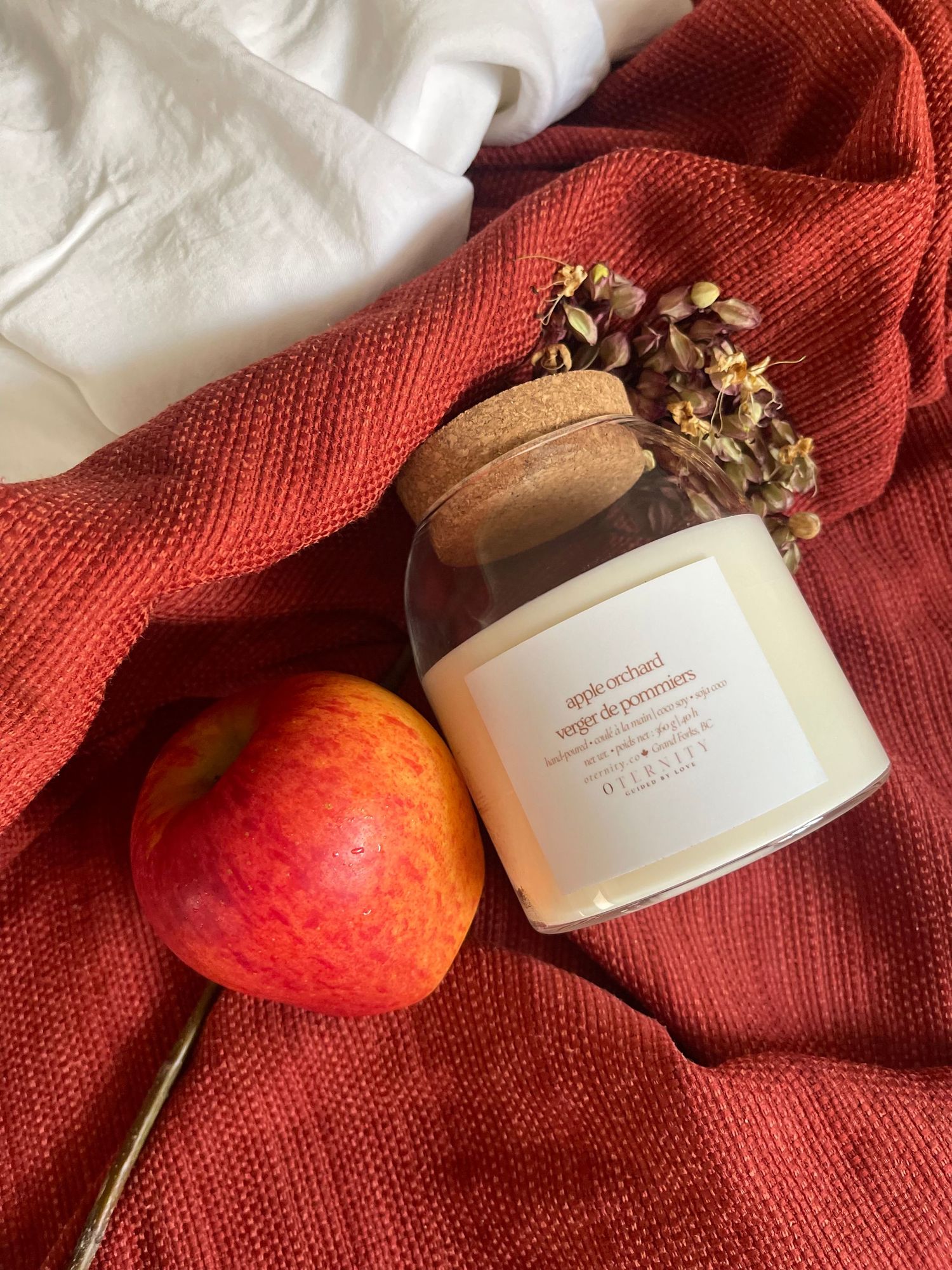 Autumn-themed candle jar with cork lid and dried flowers beside a fresh apple on red knit fabric.