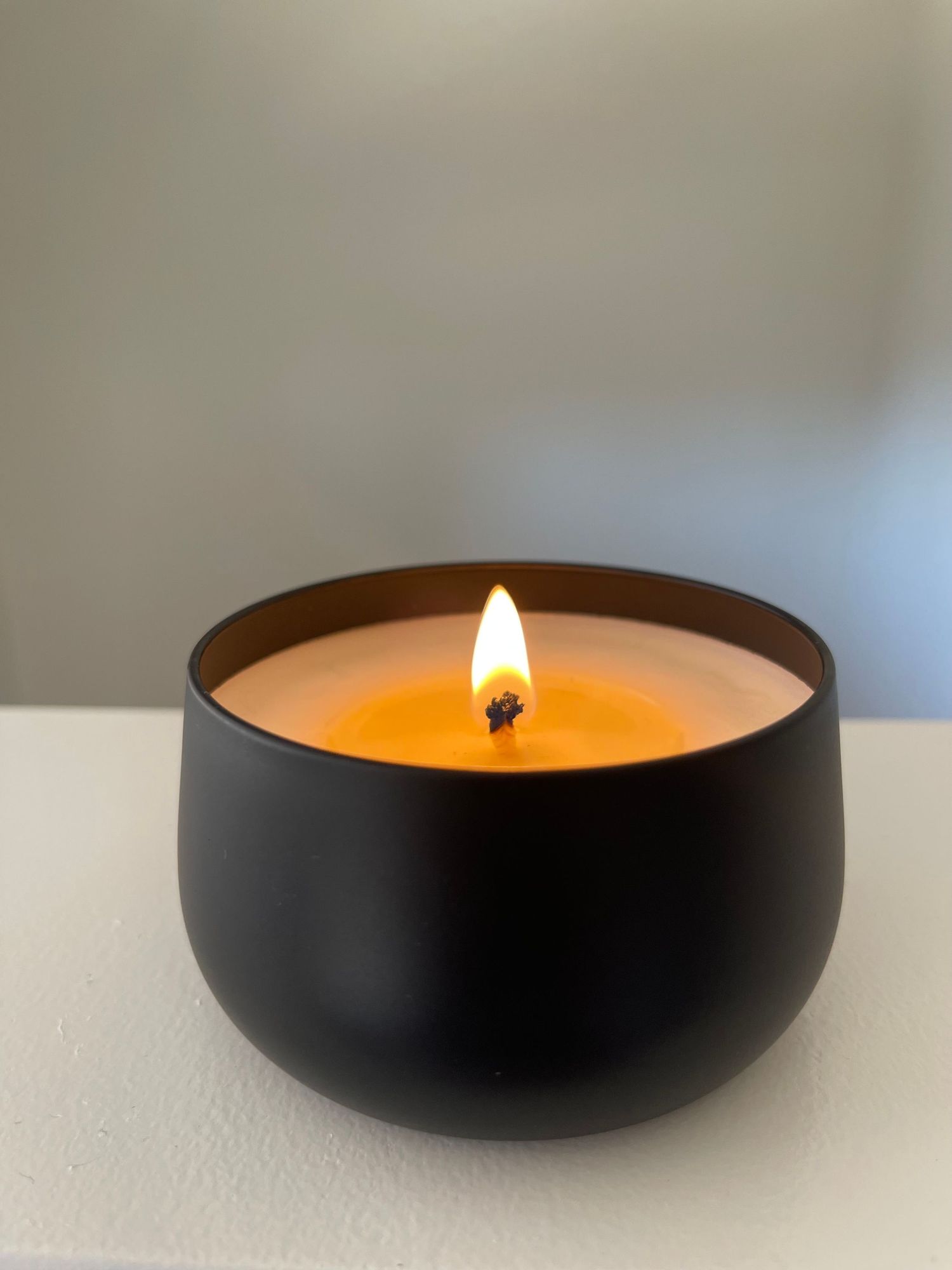 Lit black matte bowl candle with orange wax and glowing flame on white surface.