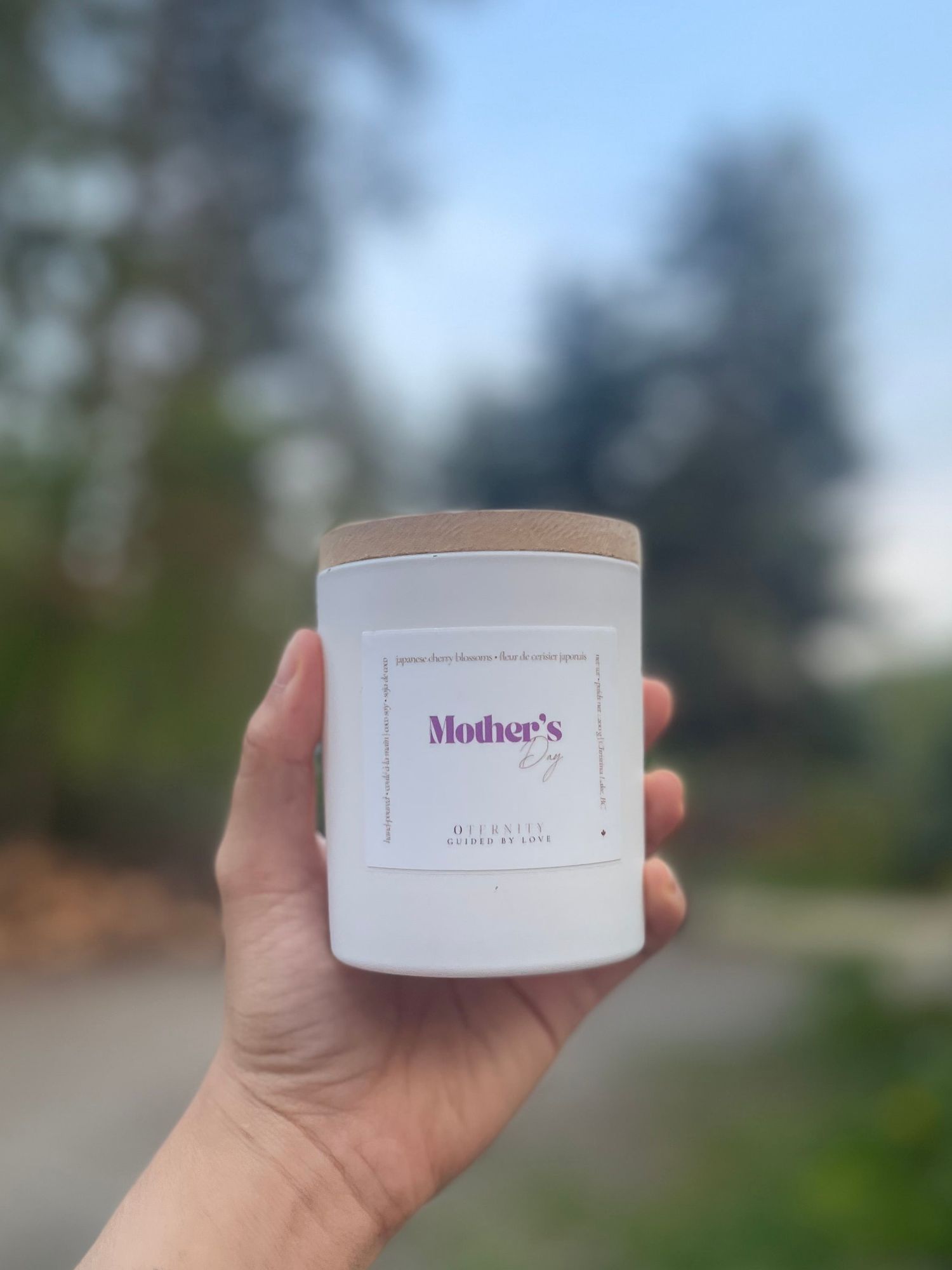Hand holding a white Mother's Day scented candle with wooden lid by Micandles, outdoors.