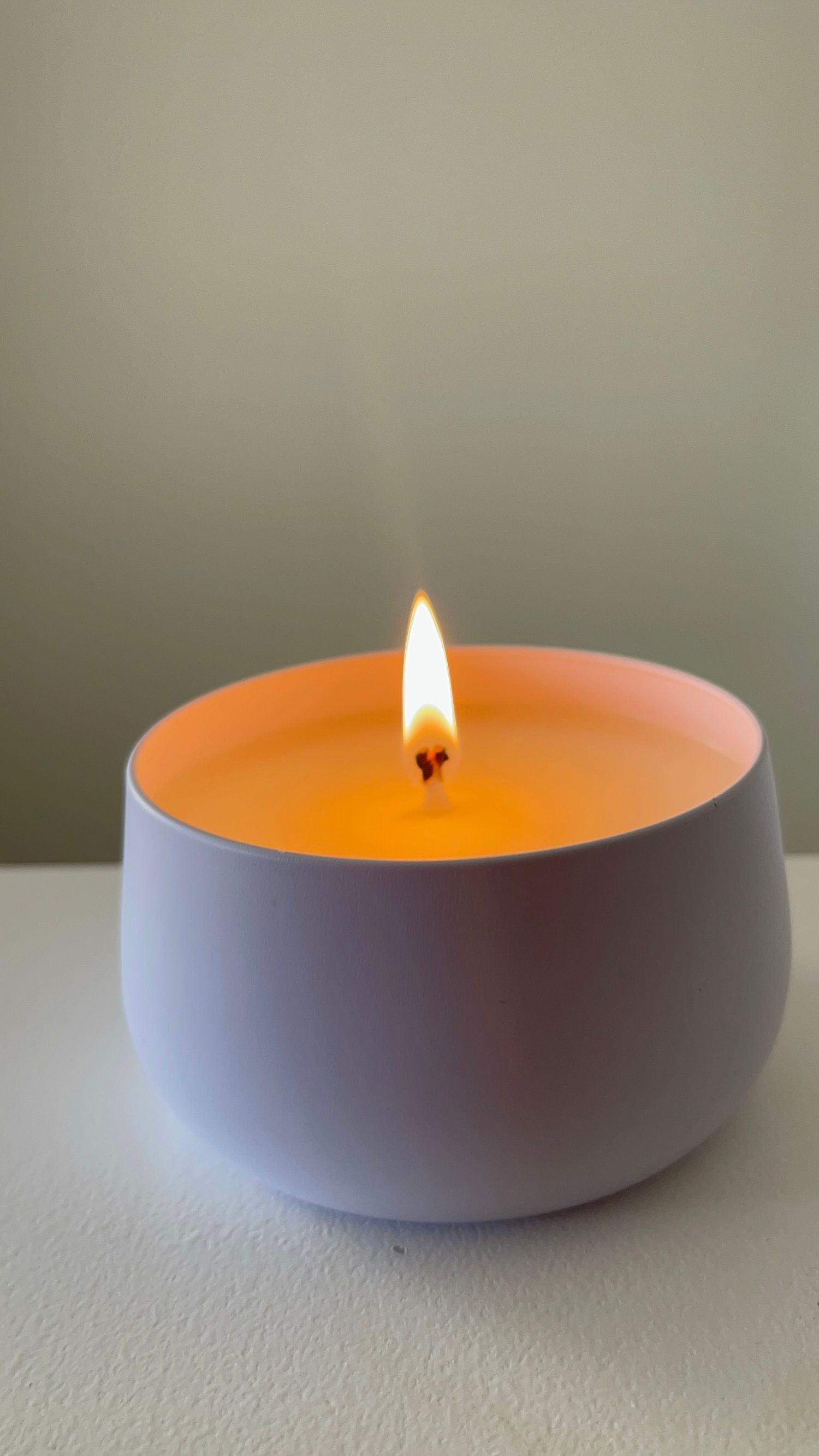 A lit candle in a round white ceramic bowl with warm orange wax on a white surface against a sage green wall.