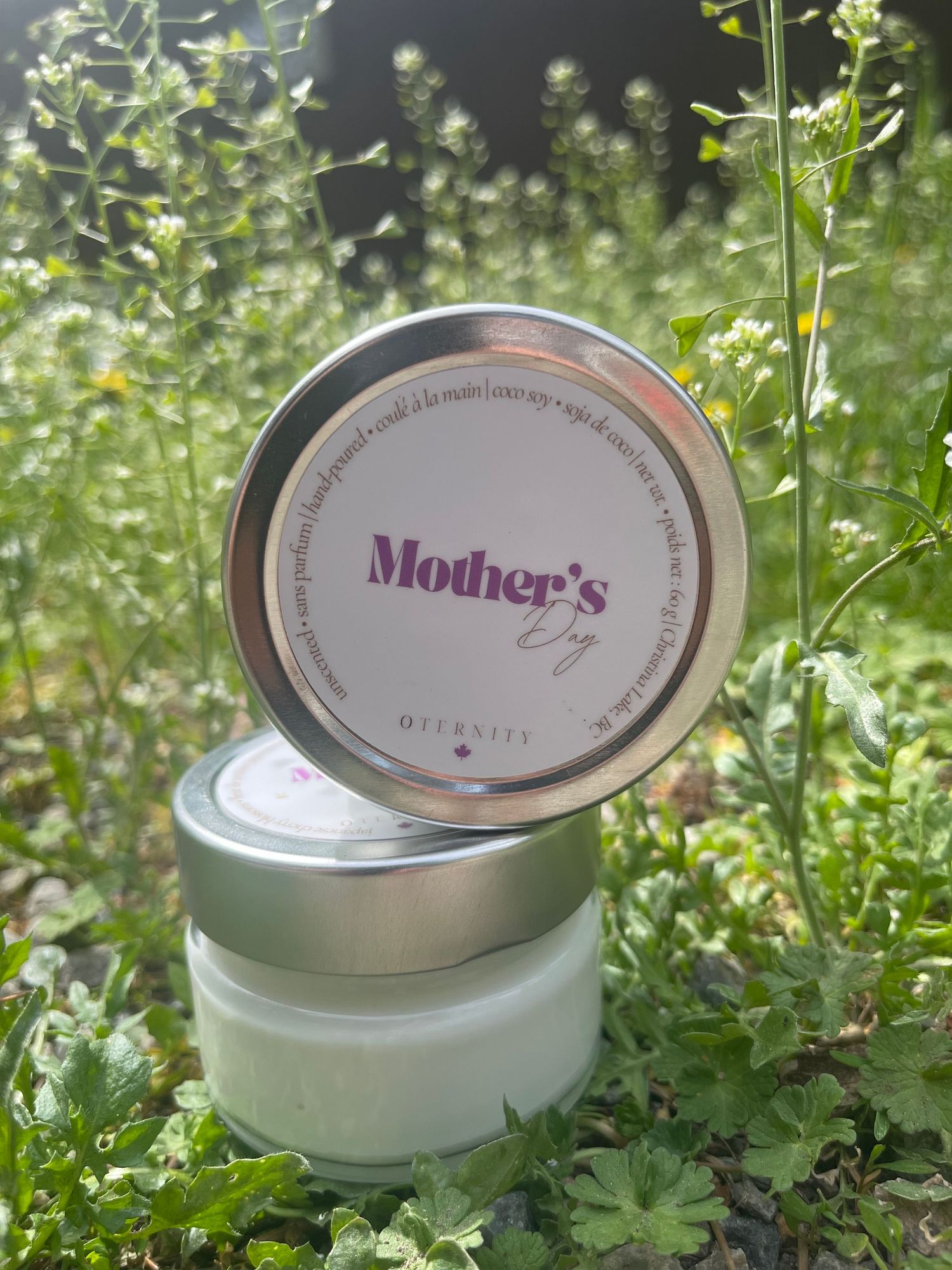 Mother's Day scented candle in silver tin displayed outdoors among green plants and wildflowers.