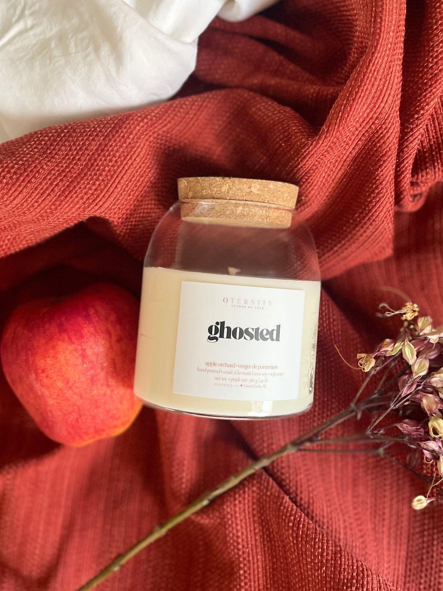 Ghosted scented candle in glass jar with cork lid, styled with a peach and dried flowers on red fabric.