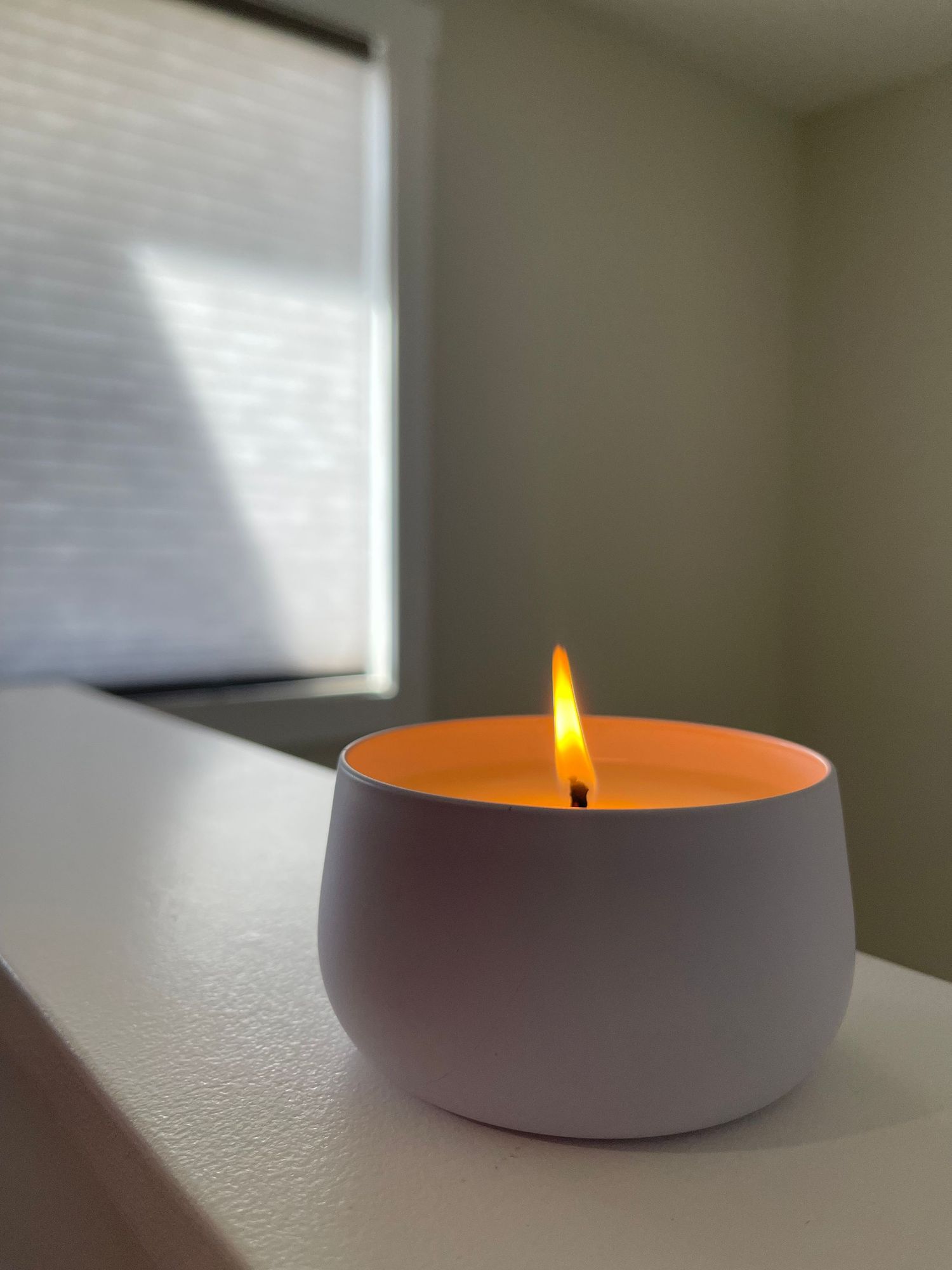 A lit white ceramic candle with an orange wax pool and glowing flame sits on a white surface near a window.