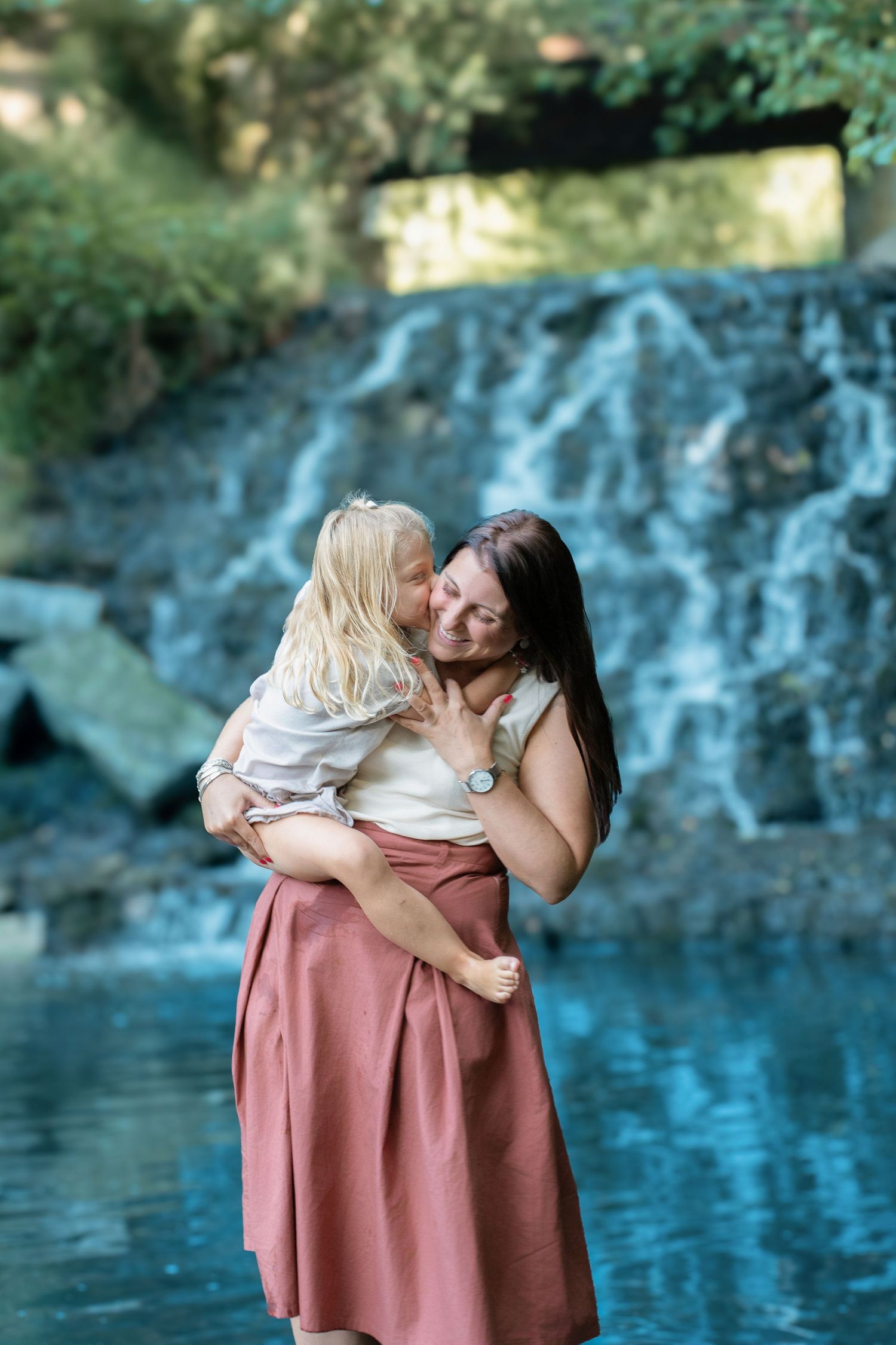A mother holds her blonde toddler near a serene waterfall, sharing a tender moment outdoors.