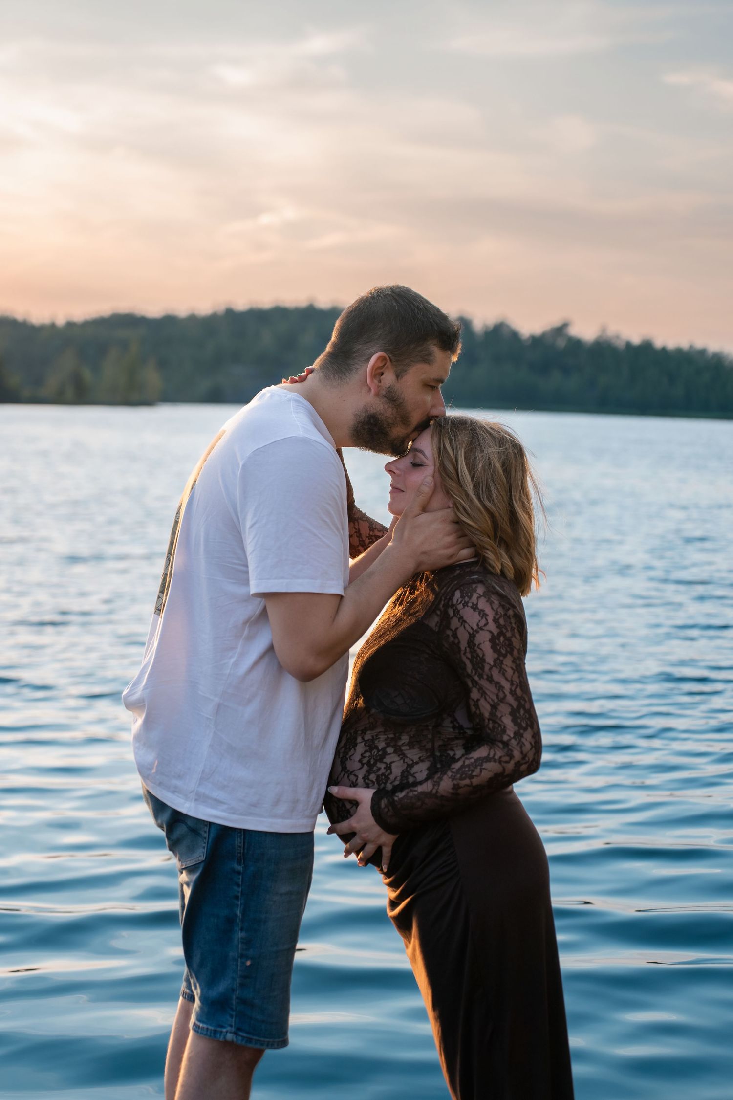 A couple shares a tender kiss by a serene lake at sunset, the woman in a black lace dress cradling her pregnant belly.