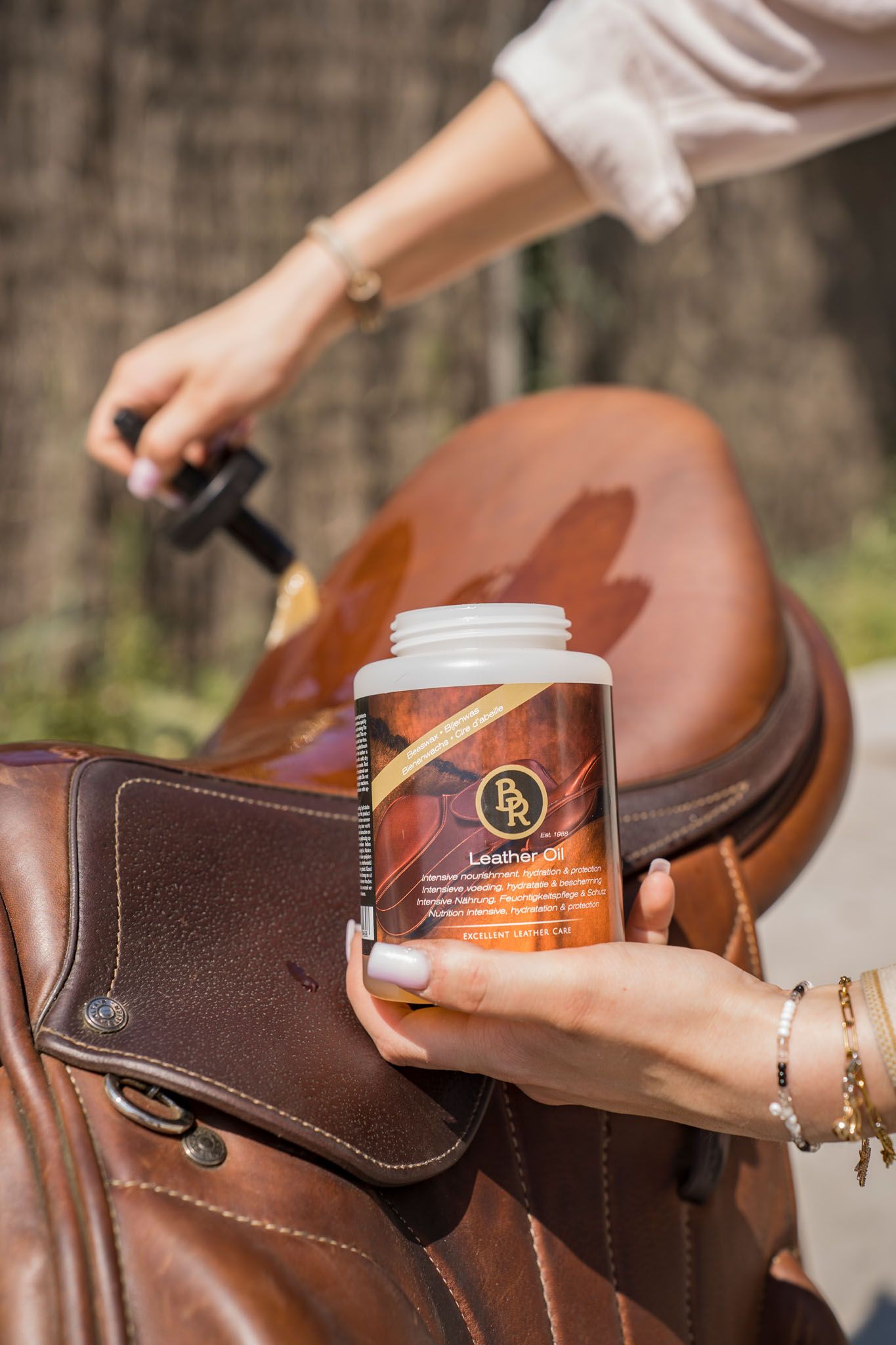Hands applying leather care products to brown equestrian saddle.