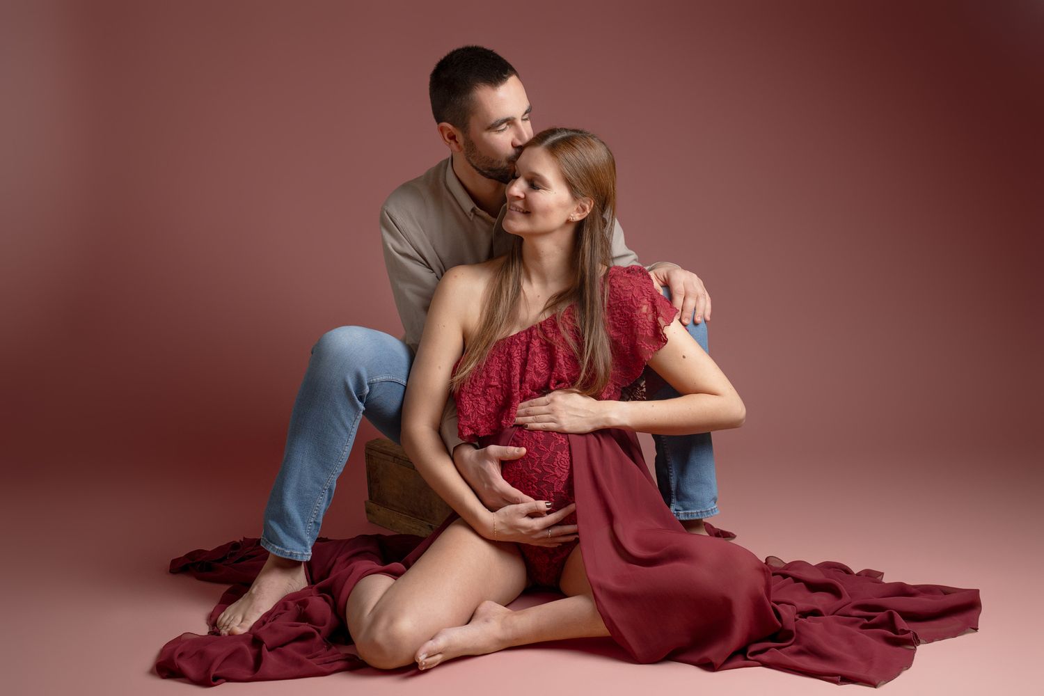 Artistic maternity photos with couple in burgundy dress against dark background.