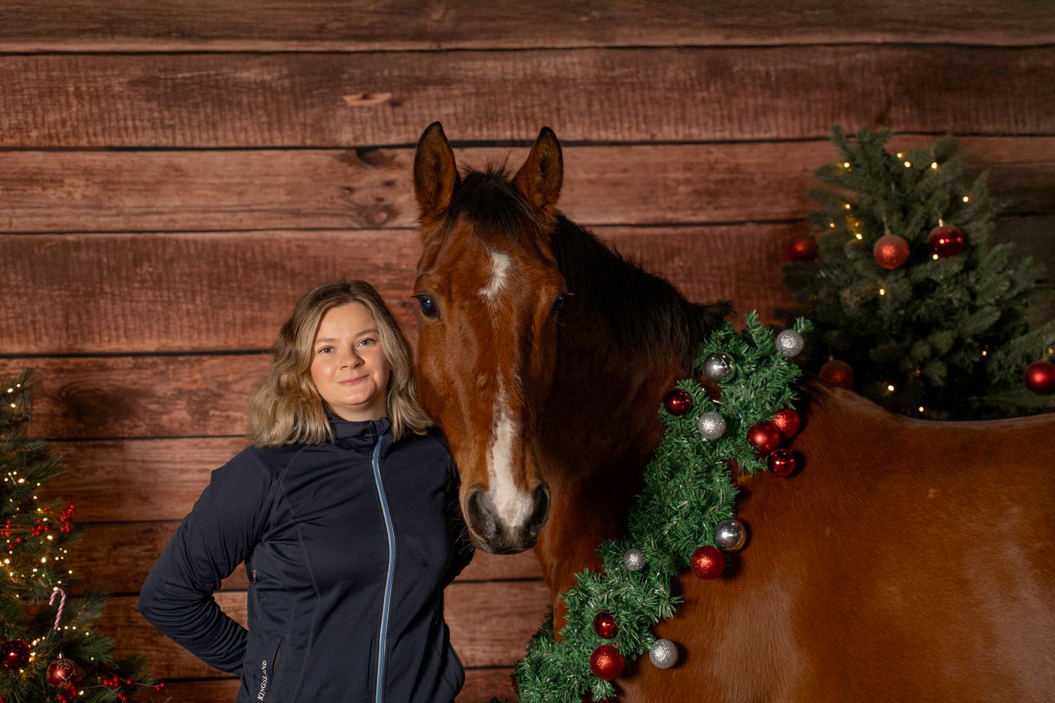 Horse and handler pose by Christmas decorations in rustic barn setting.
