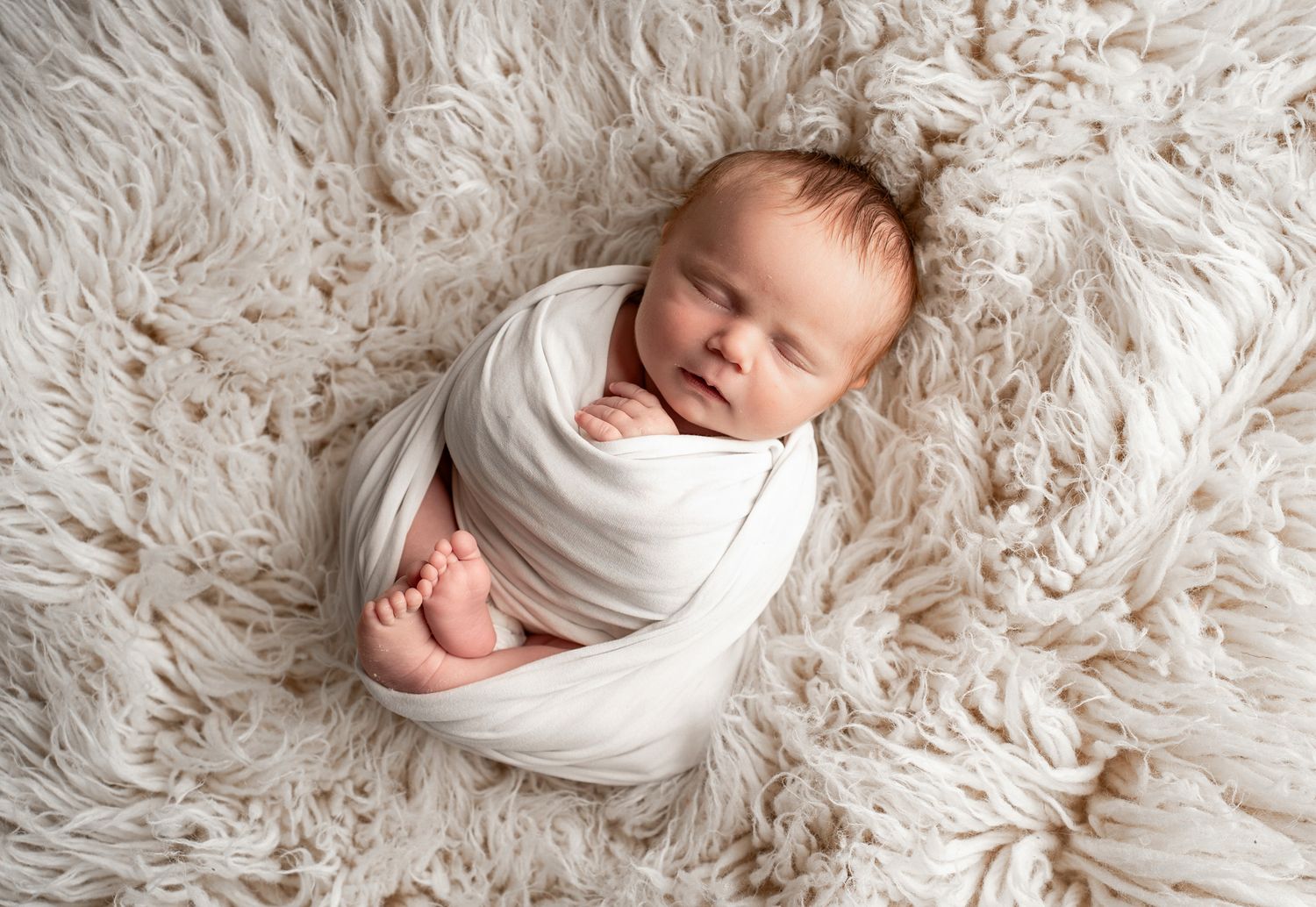 Sleeping newborn wrapped in white swaddle on plush cream-colored fur backdrop.