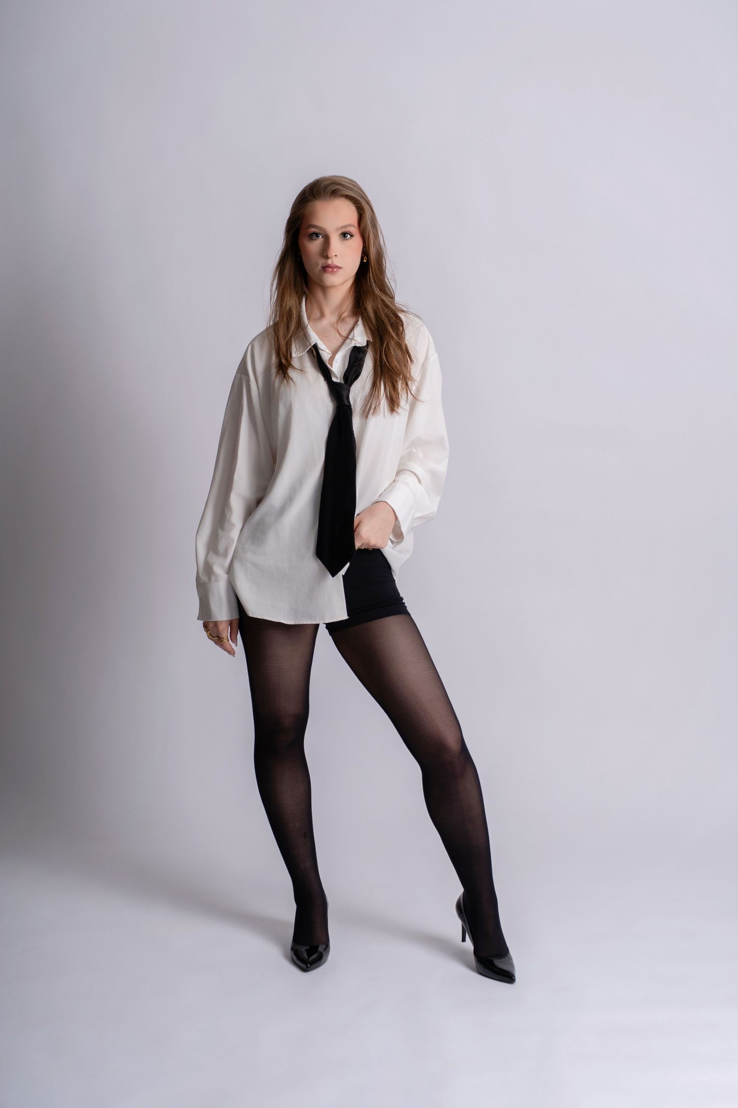 Young woman in oversized white shirt, black tie, shorts, sheer tights, and heels posing against gray background.